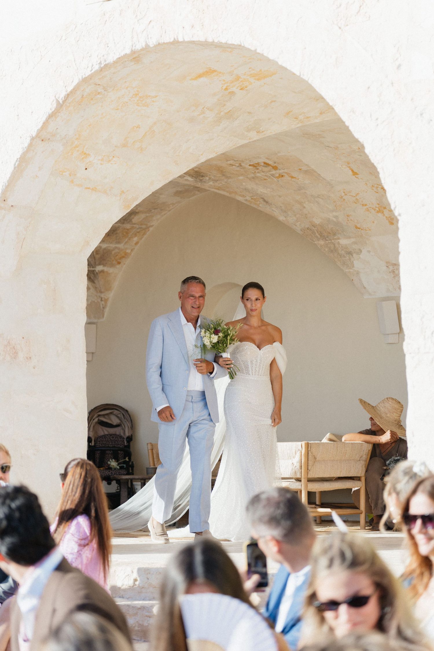 A father in a light blue suit walks his bride in an off-shoulder gown through a stone archway as wedding guests watch.