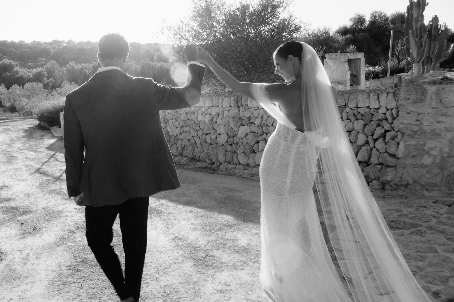 Black and white photo of a bride in a flowing veil and groom walking hand in hand beside a stone wall.