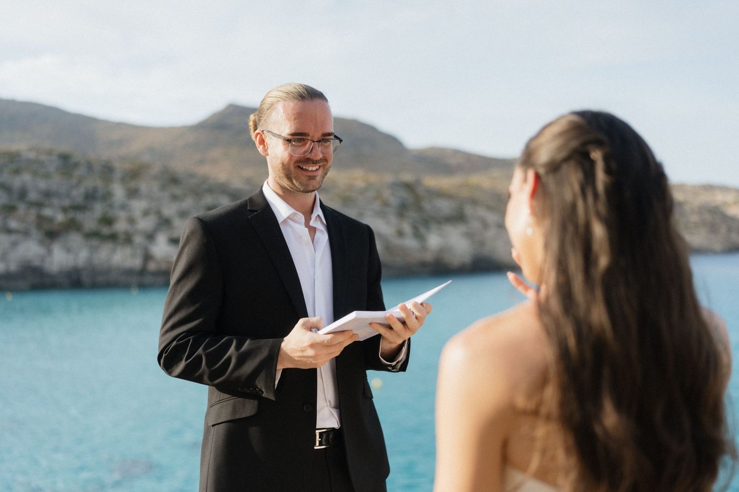 Smiling officiant in a dark suit and glasses reads from a paper to a woman with long hair at a seaside wedding ceremony with rocky coast.