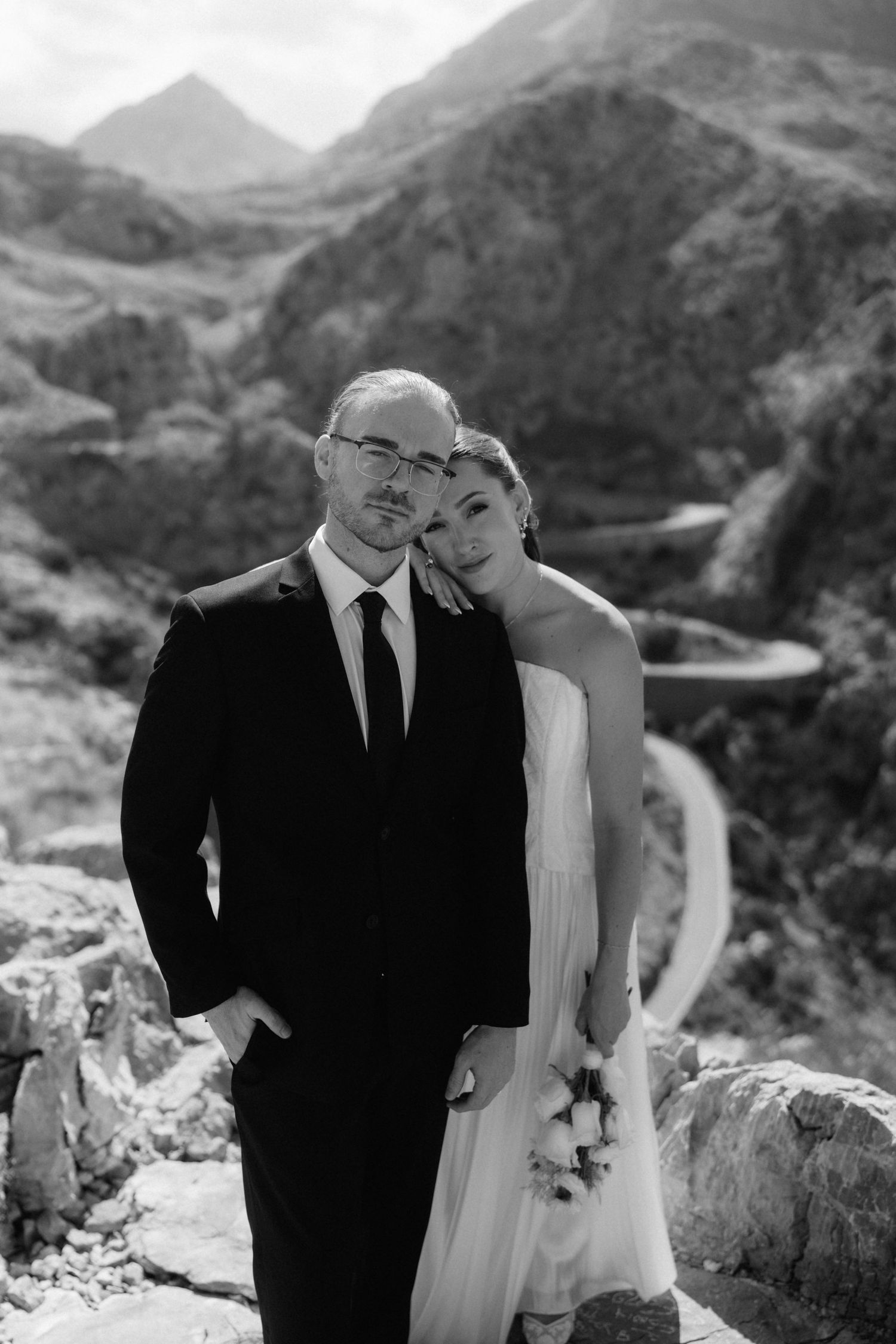 Black and white wedding portrait of a bride leaning on a groom in a suit and glasses, with rugged mountains and a winding road behind.