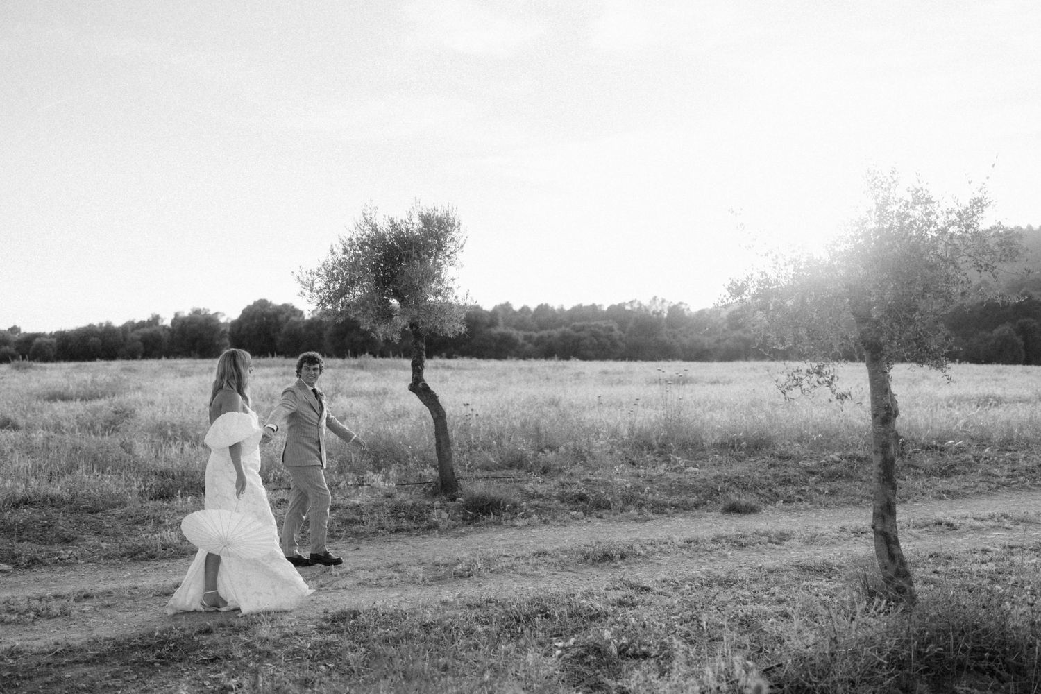 Black and white photo of a bride and groom walking through a sunlit field with trees in a romantic outdoor setting.