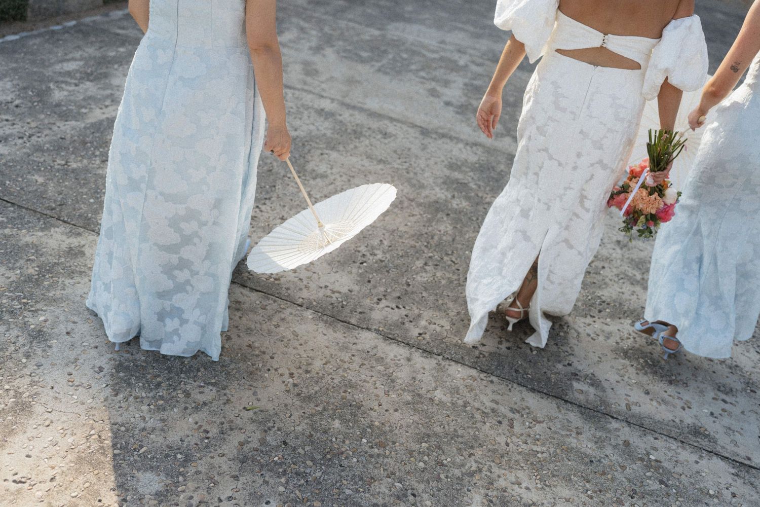 Two brides in white lace gowns walking, one holding parasol, other carrying bridal bouquet.