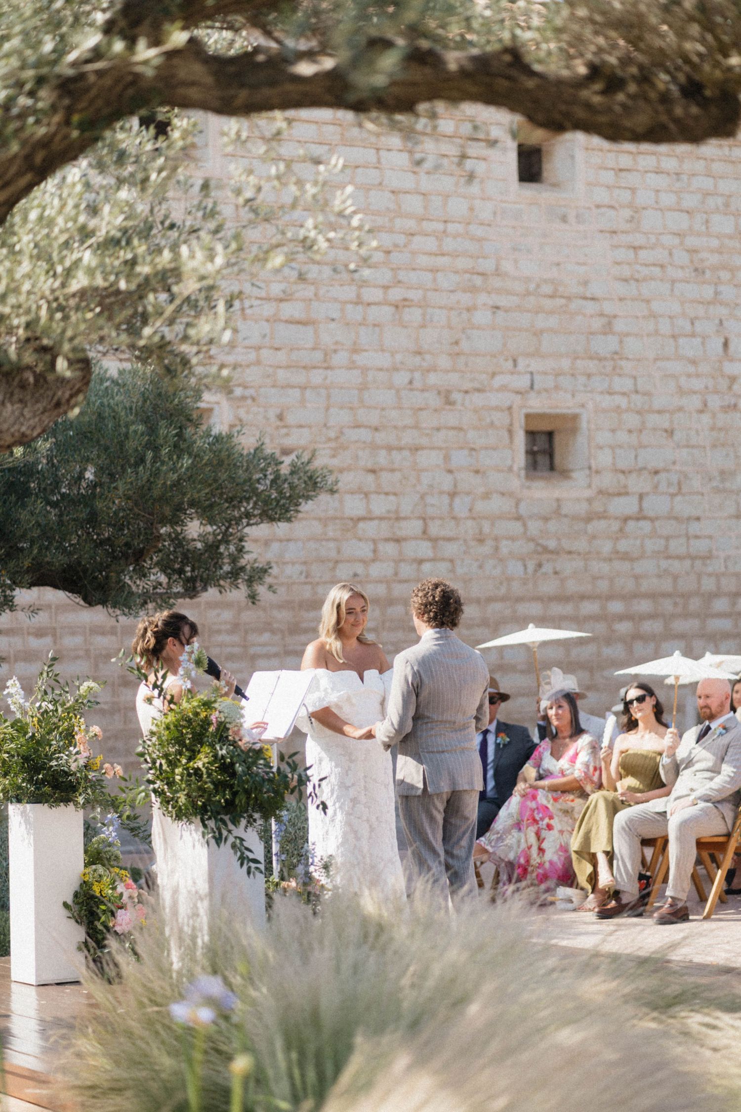 Bride and groom exchange vows at outdoor ceremony beside stone building with olive trees and floral arrangements.
