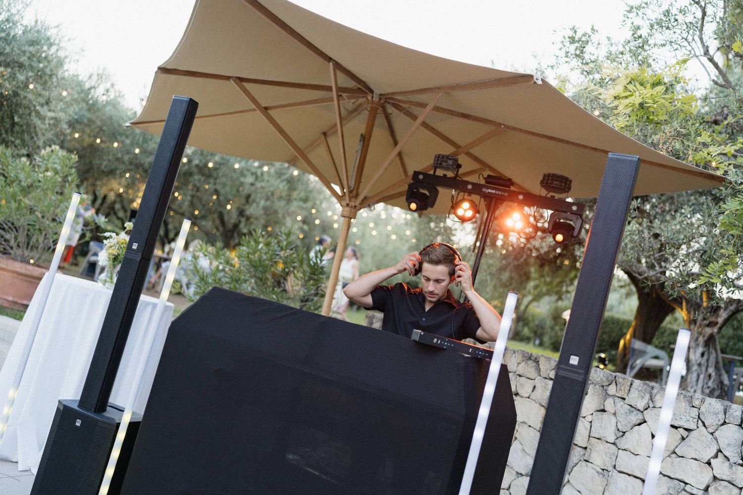 DJ performing under a large umbrella at an outdoor evening event surrounded by olive trees and string lights.