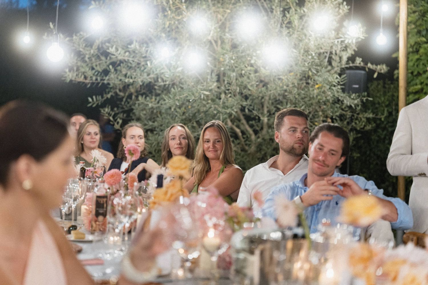 Guests enjoying an outdoor wedding reception dinner under string lights surrounded by lush greenery and candlelit tables.