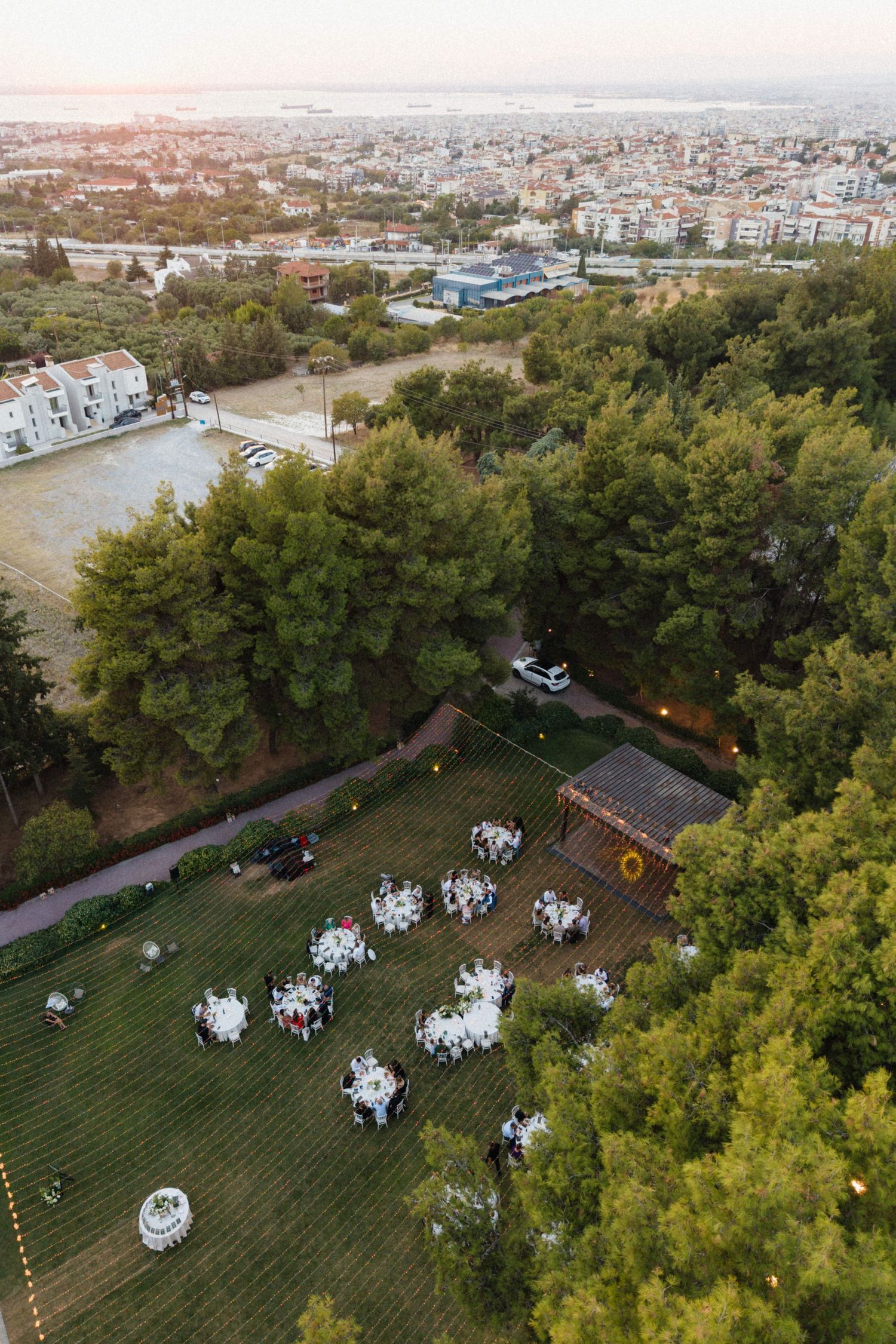 Aerial view of outdoor wedding reception with round white tables on green lawn surrounded by trees at dusk.