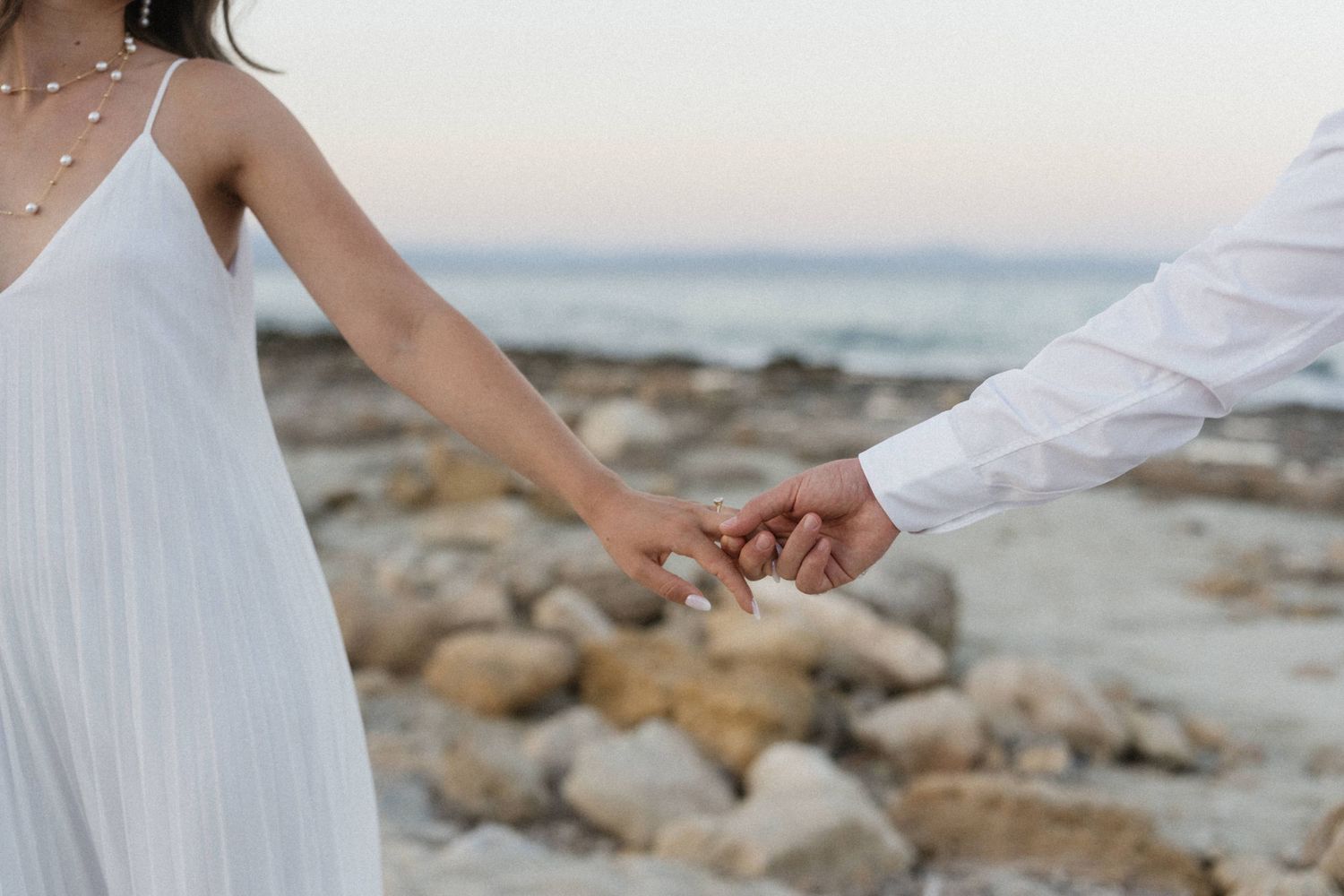 Close-up of bride and groom holding hands near rocky ocean shore, bride in white dress with pearl necklace.