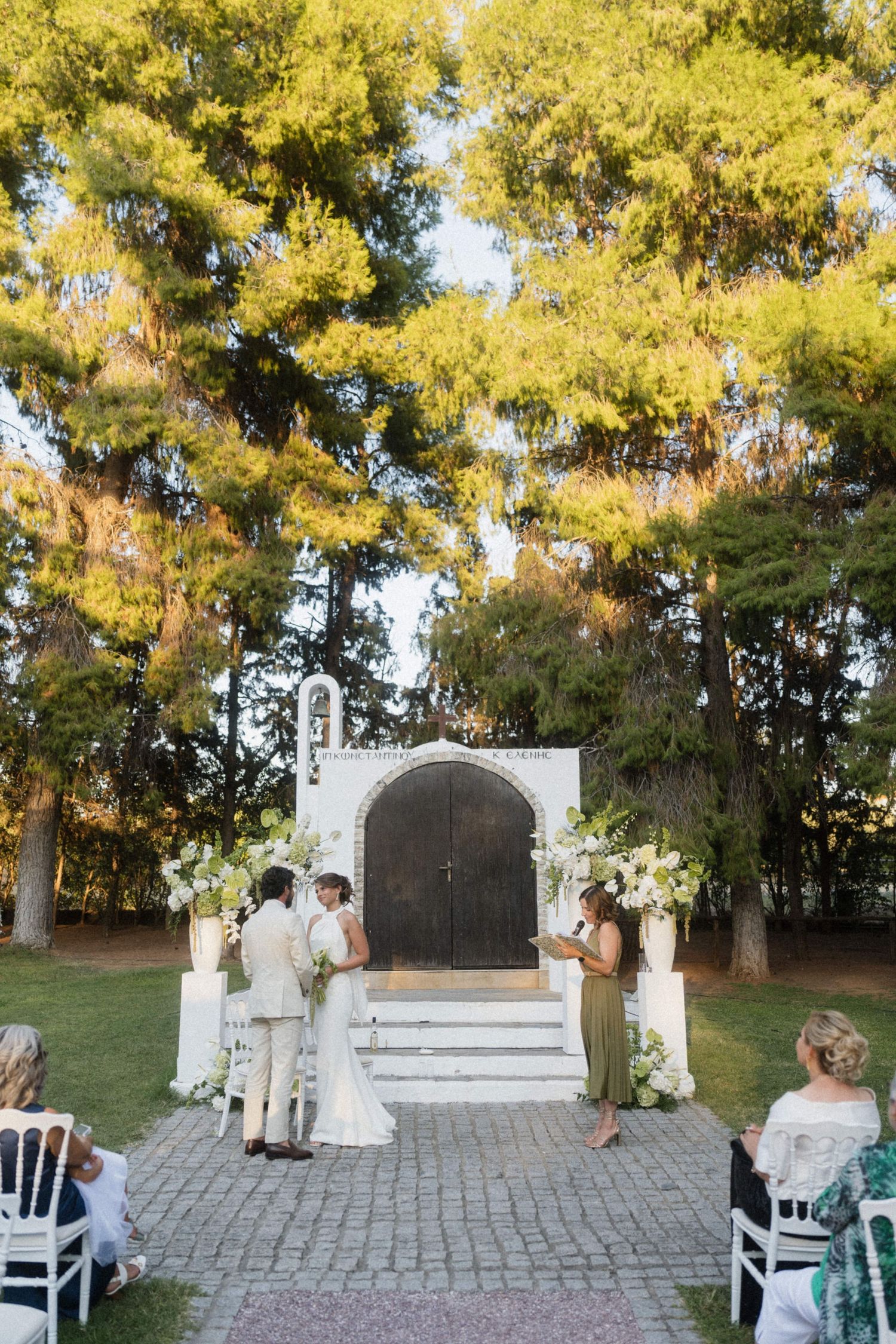Outdoor wedding ceremony at ornate white chapel doors framed by tall trees with white floral arrangements.
