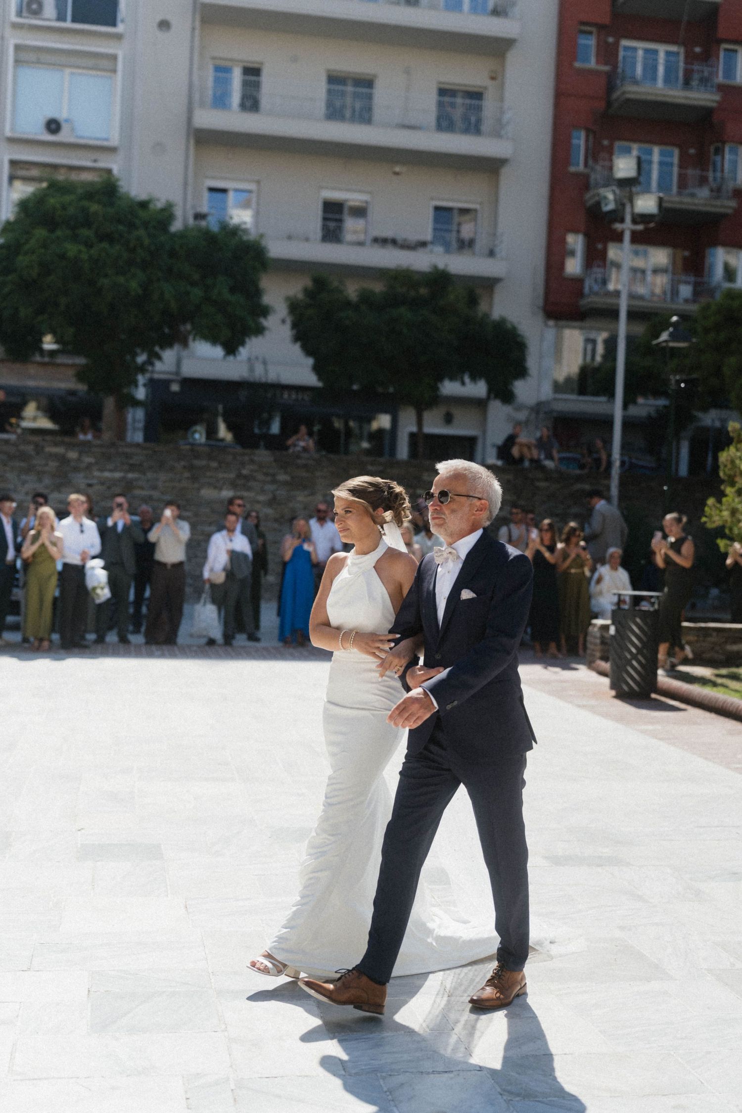 Bride in white halter gown walks with escort across sunlit plaza with wedding guests watching.