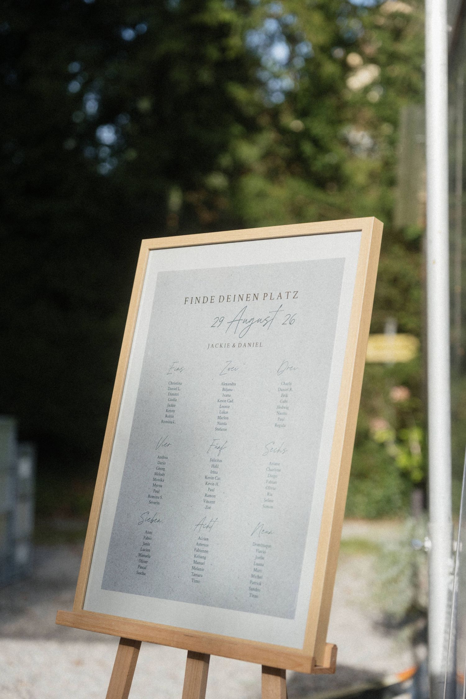 Wooden framed wedding seating chart on easel displayed outdoors among green trees at garden wedding.