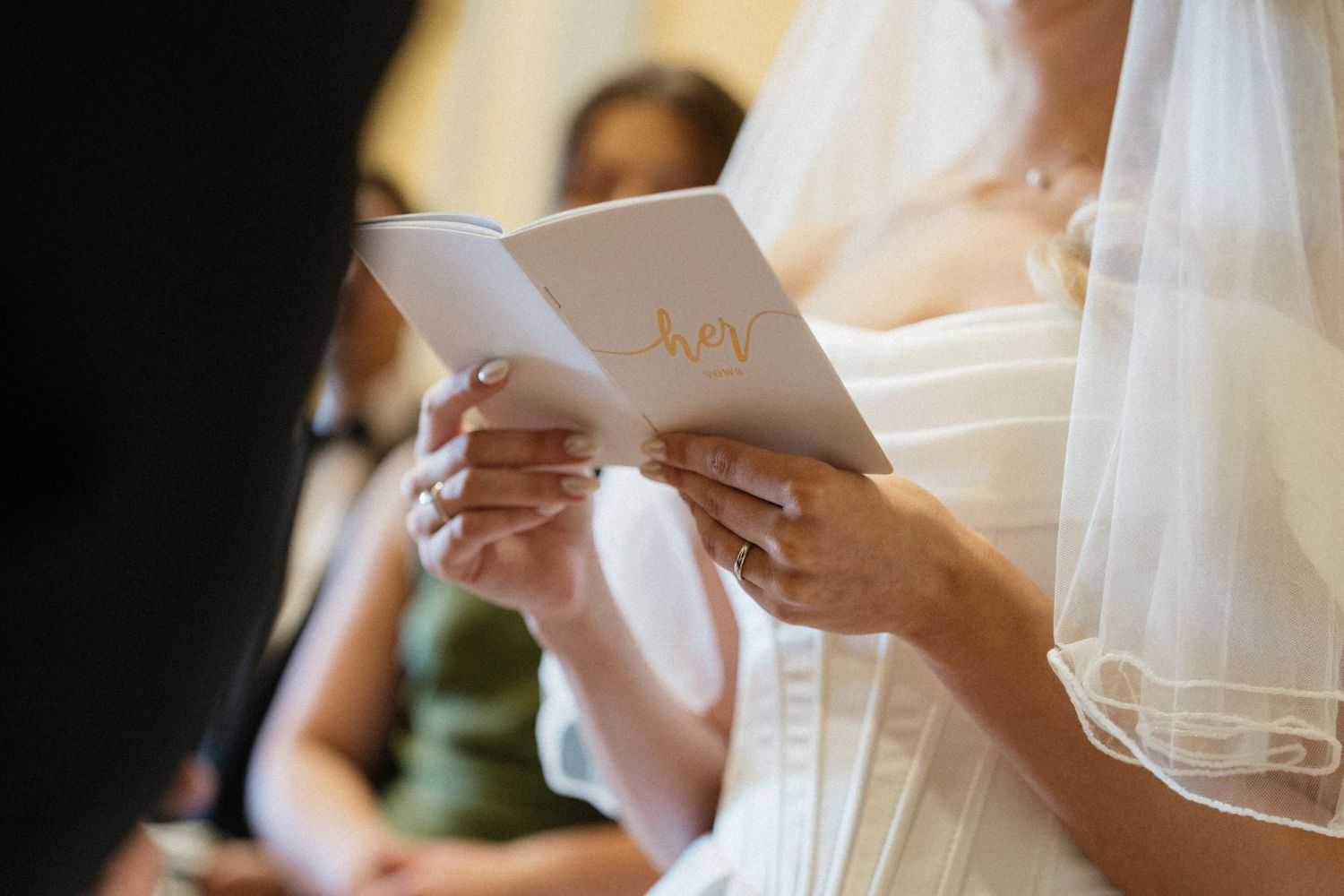 Close-up of bride holding wedding program booklet with delicate veil and manicured nails during ceremony.