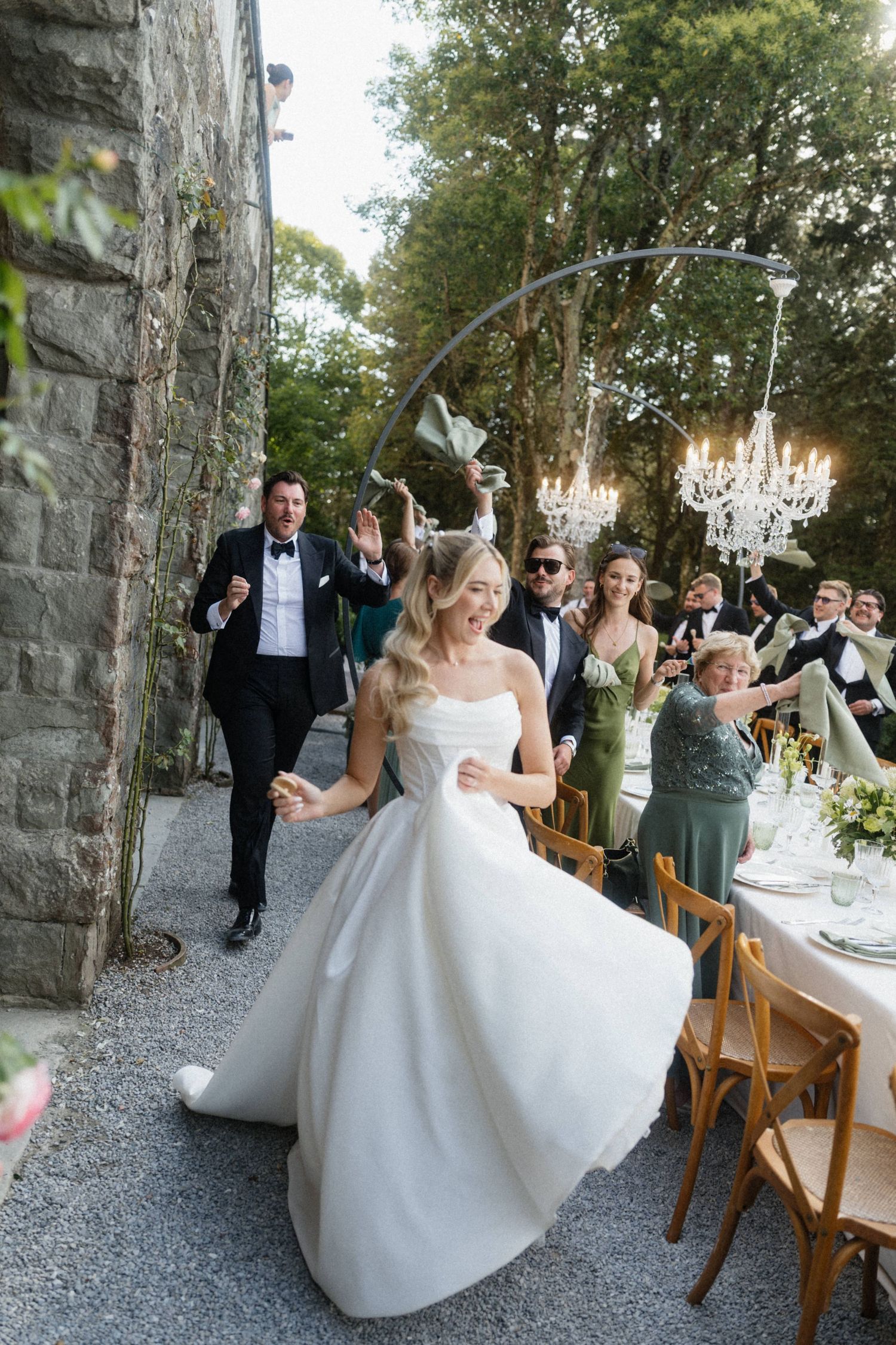 Bride in white ball gown leads joyful wedding party entrance along outdoor reception table at Italian villa.