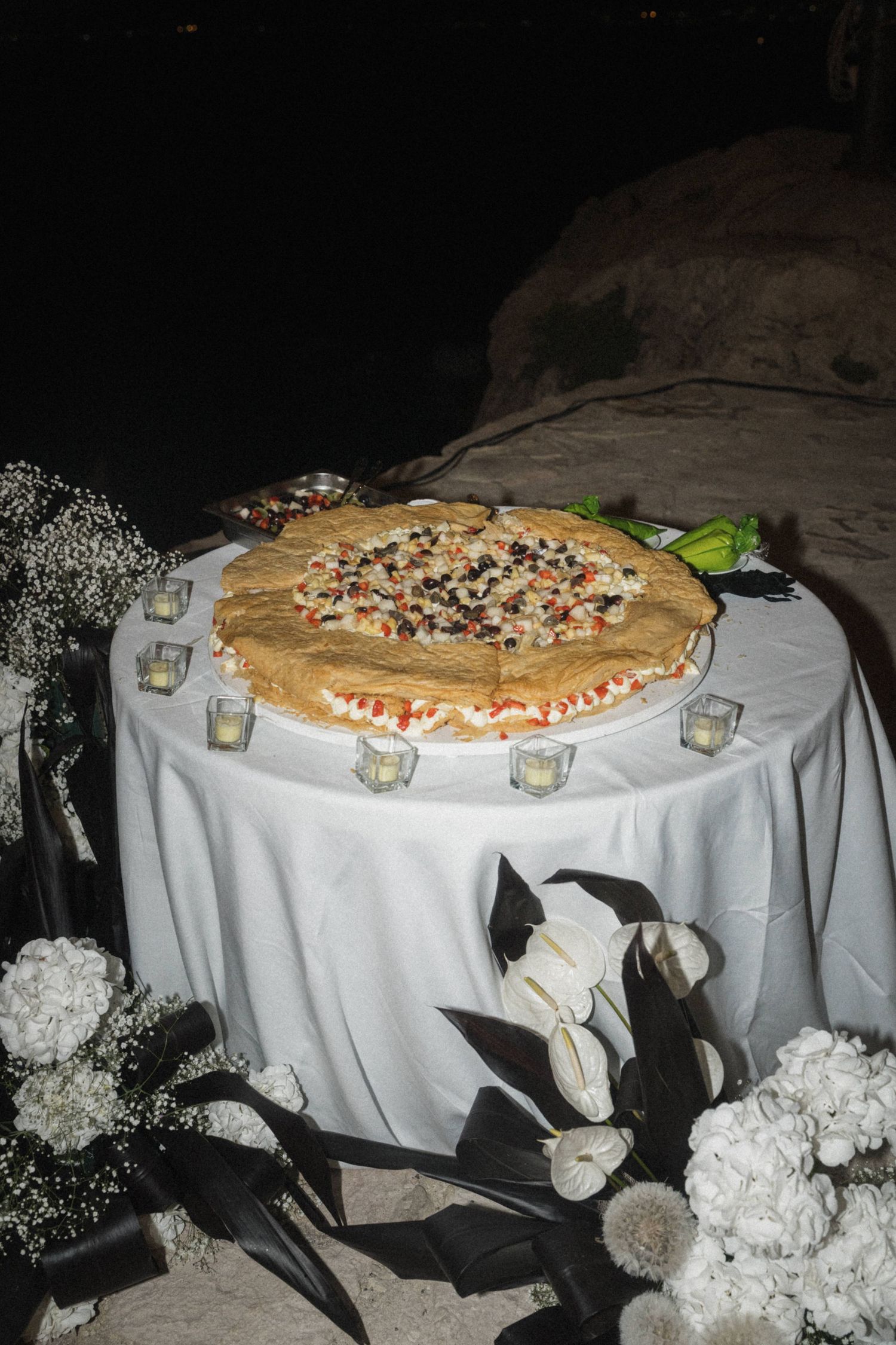 Large decorated wedding pizza cake on white draped table with white floral arrangements at night.