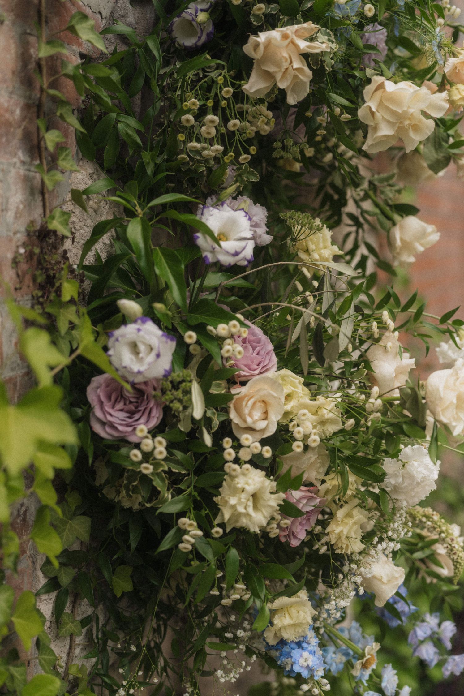 Lush floral arrangement with purple, white, and cream roses and greenery decorating a rustic brick wall.