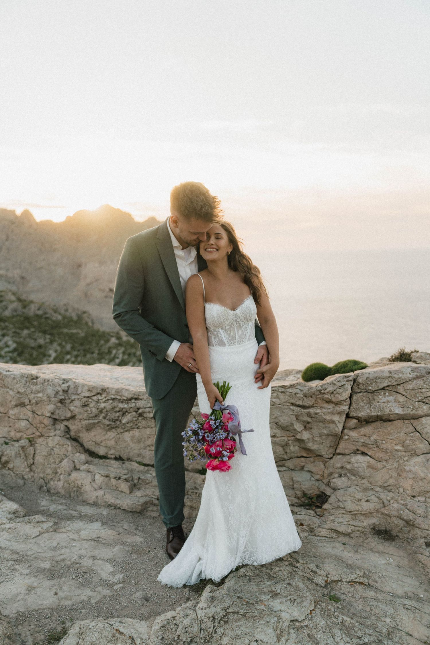 Wedding couple pose at sunset on stone wall overlooking the sea, bride holding vibrant pink bouquet in fitted lace gown.