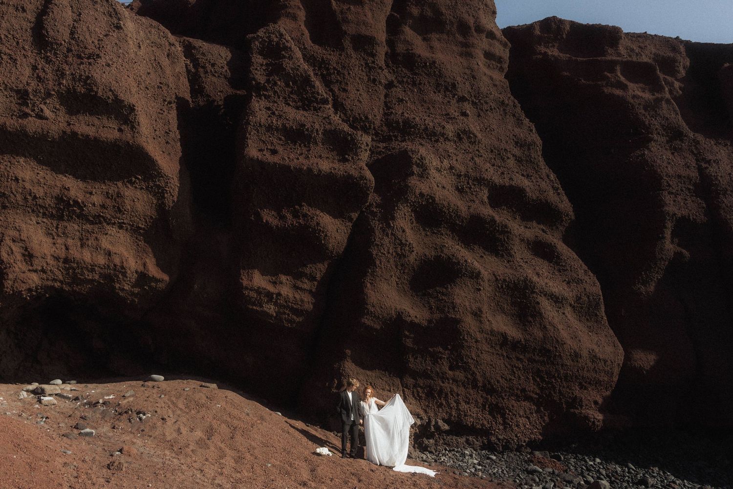 Bride and groom posing before dramatic red volcanic rock formations on their wedding day.