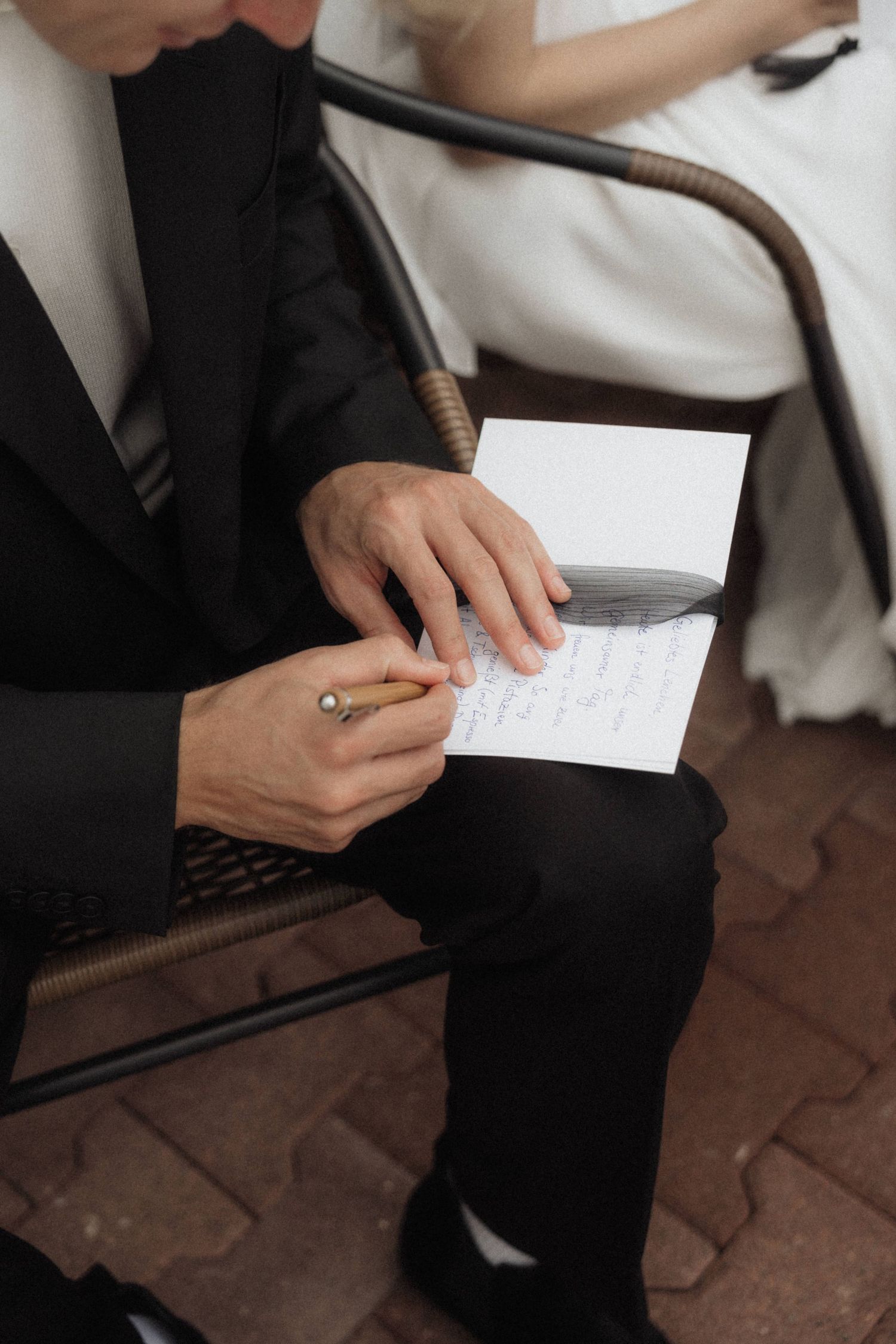 Groom in black suit reads handwritten wedding vows from a card while seated next to his bride.