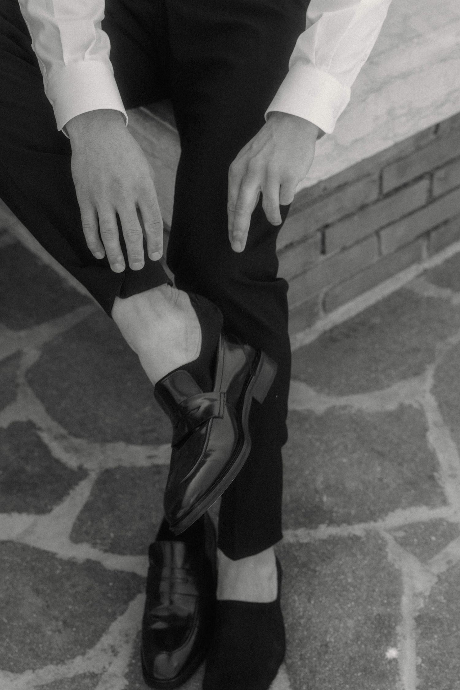 Man in white shirt and black trousers putting on shiny black dress shoes on a stone patio.