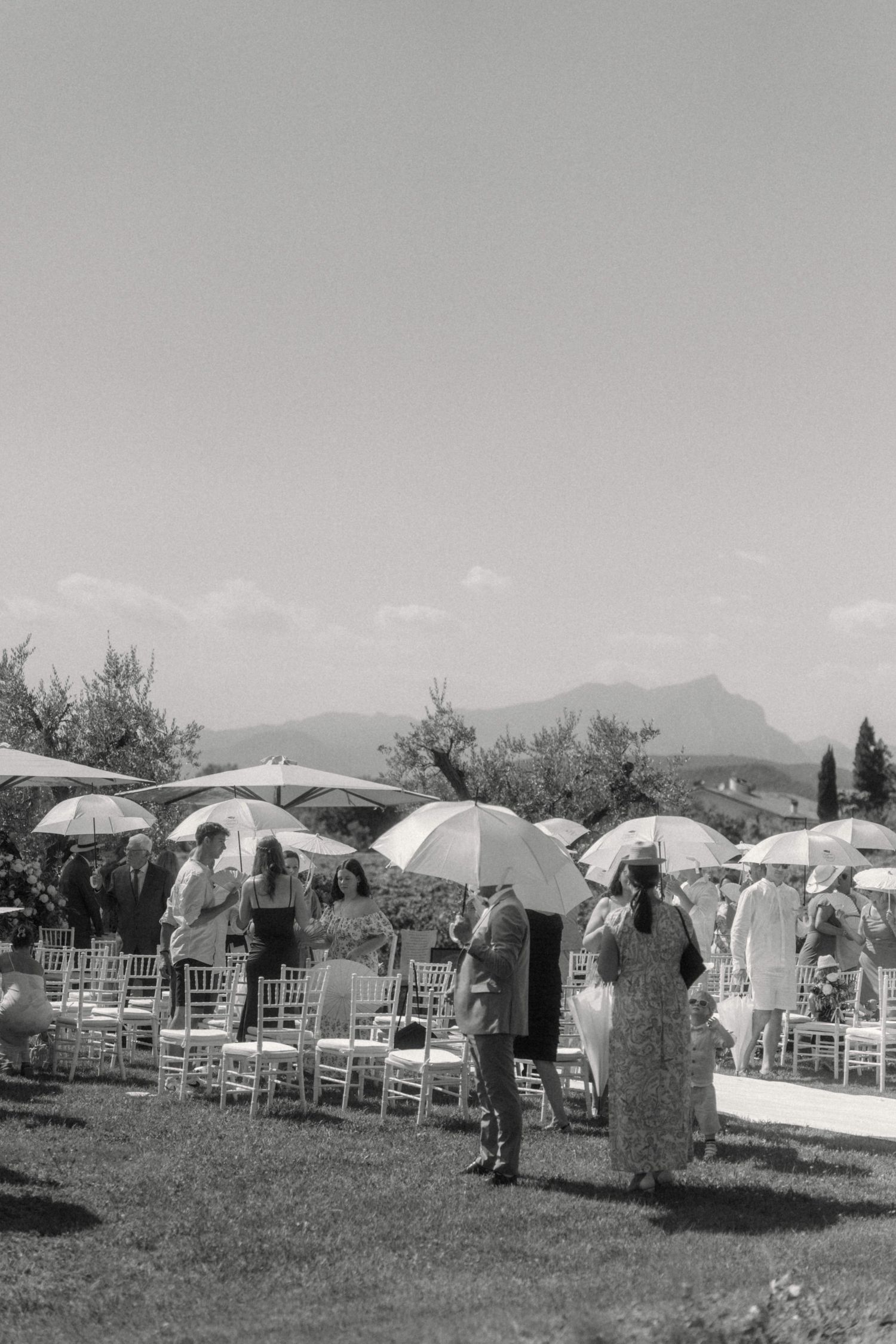 Black and white photo of outdoor wedding ceremony with guests holding parasols and mountain views.