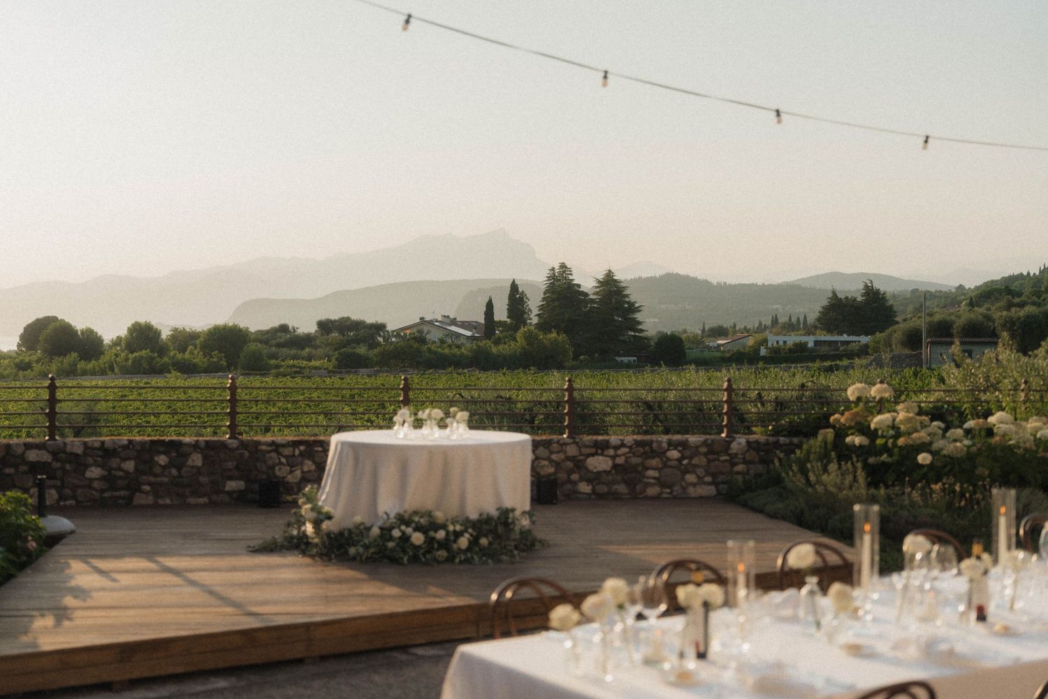Romantic vineyard wedding reception setup on wooden deck with mountain views and string lights at dusk.