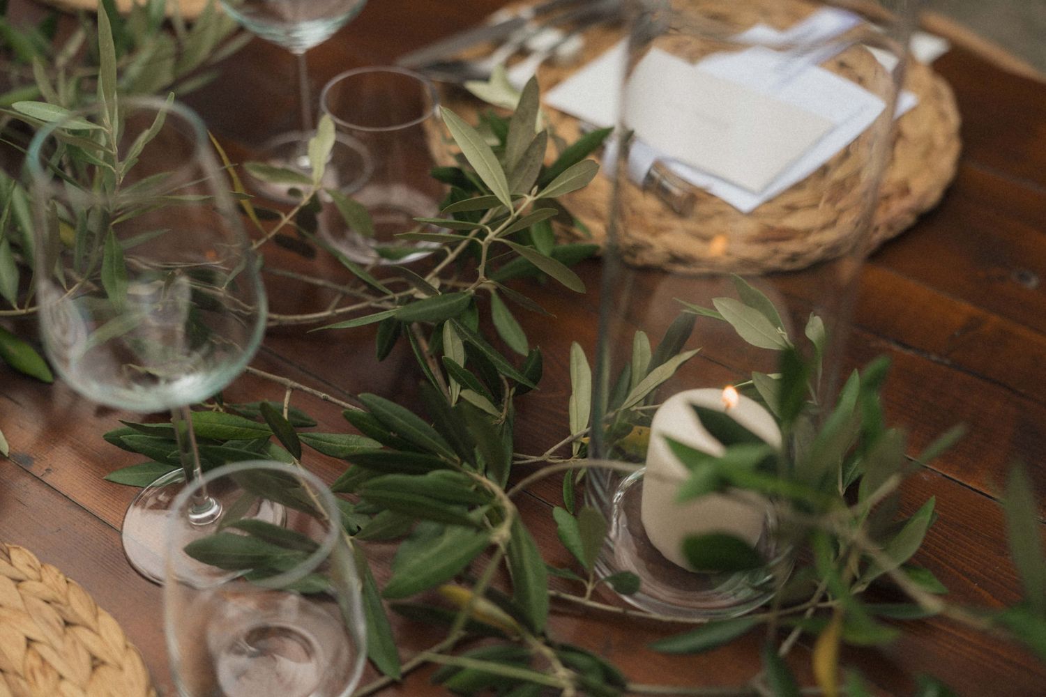 Rustic wedding table setting with olive branch greenery, clear glasses, and wicker charger plates.
