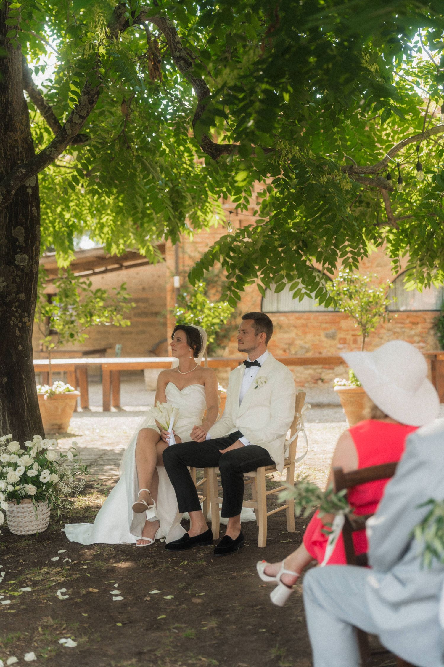 Bride and groom seated together under green tree canopy during outdoor wedding ceremony.