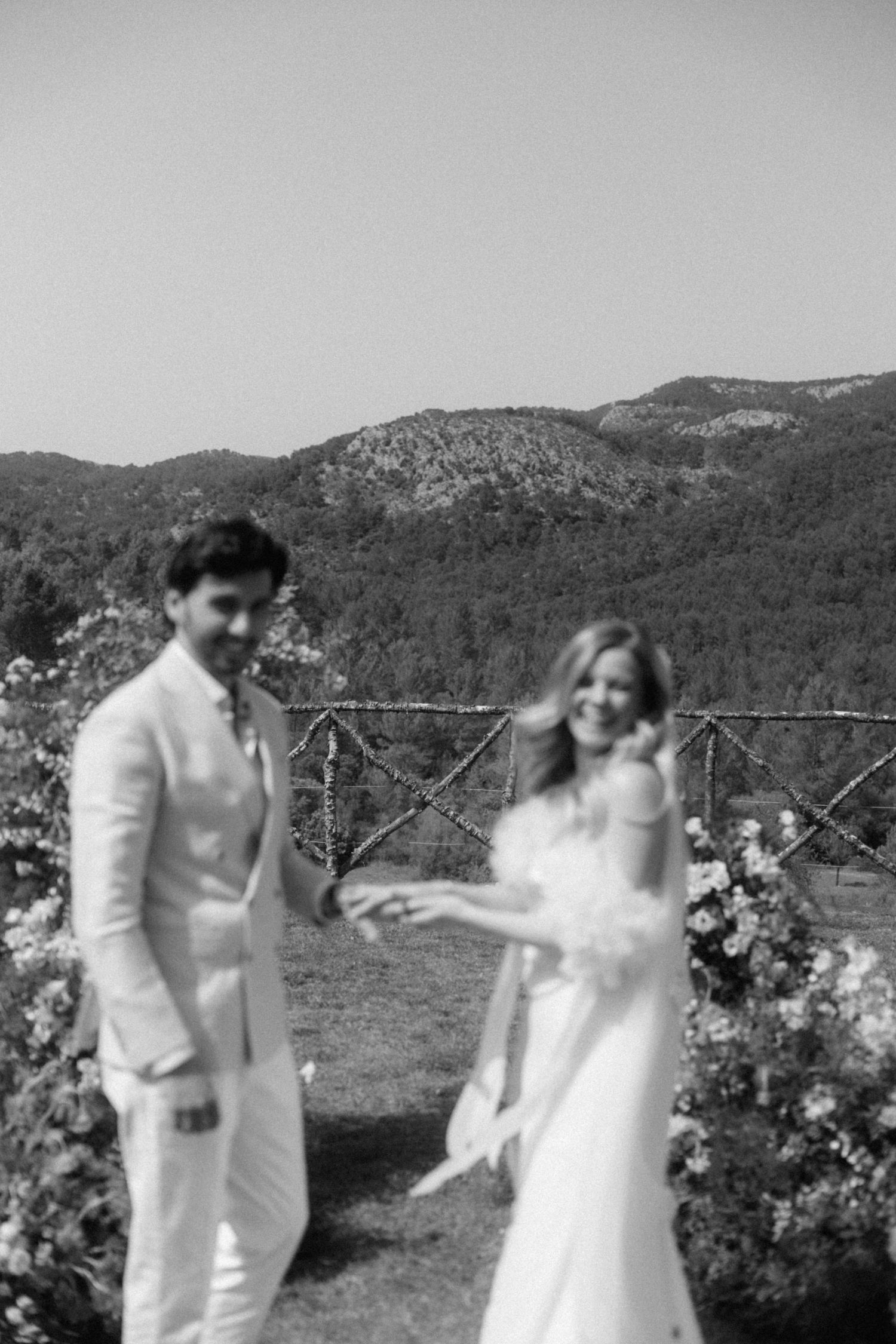 Black and white photo of bride and groom holding hands at outdoor wedding ceremony with mountain views.