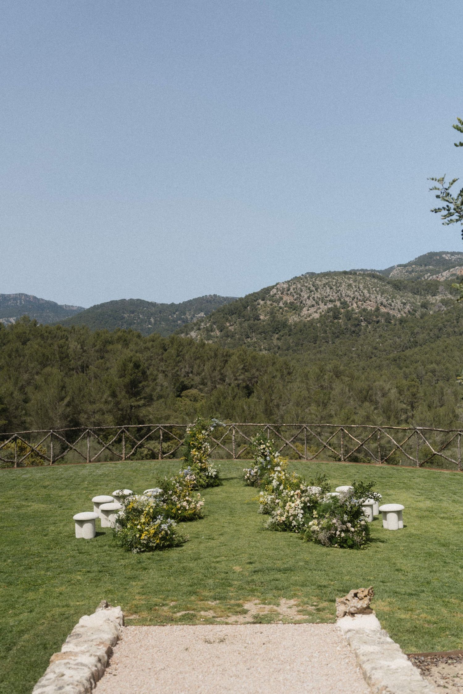 Outdoor wedding ceremony setup on green lawn with floral arrangements, white stools, and scenic mountain views under clear blue sky.