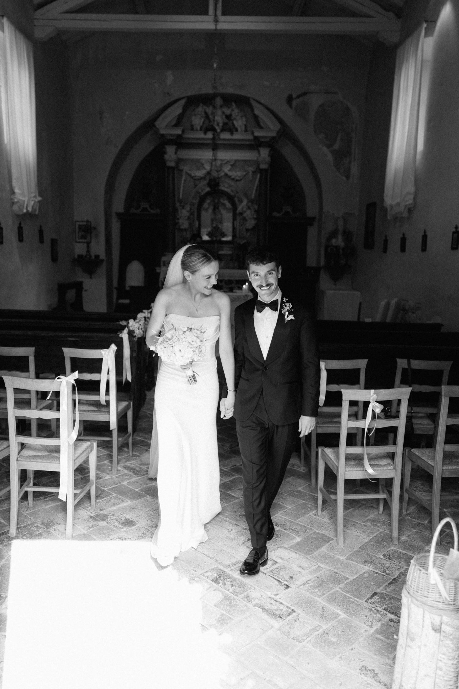 Newlyweds walk hand-in-hand down the aisle of a charming rustic stone chapel after ceremony.
