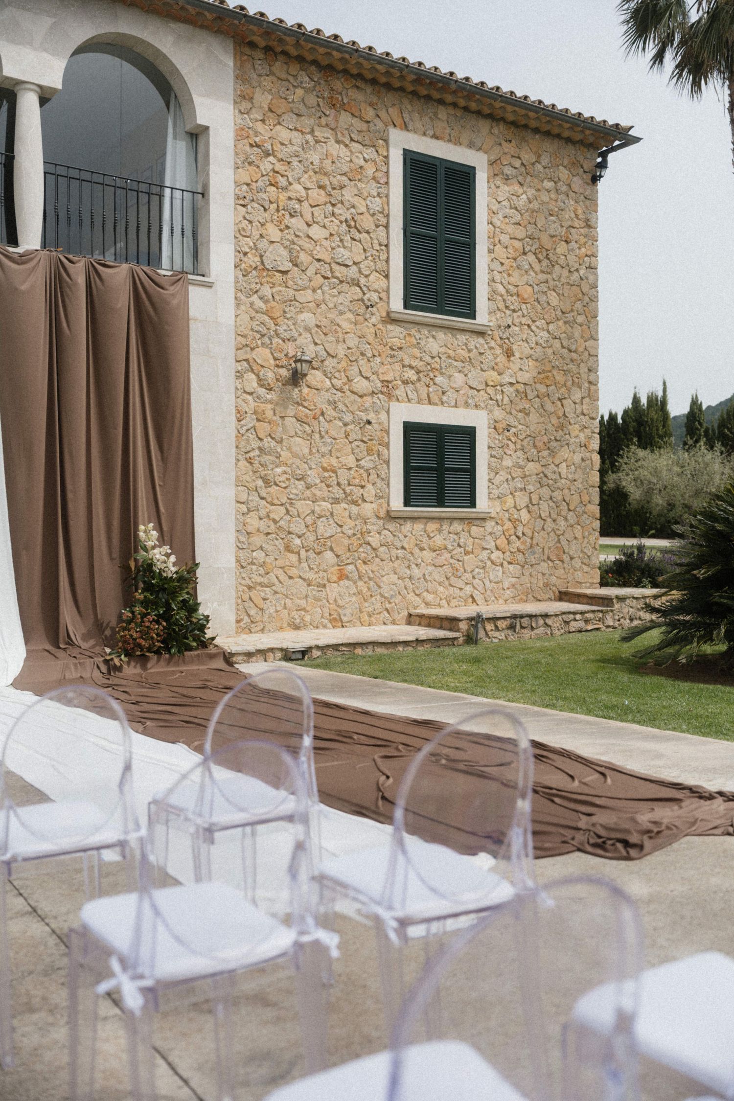 Elegant outdoor wedding ceremony setup with ghost chairs and brown fabric runner before a rustic stone villa.