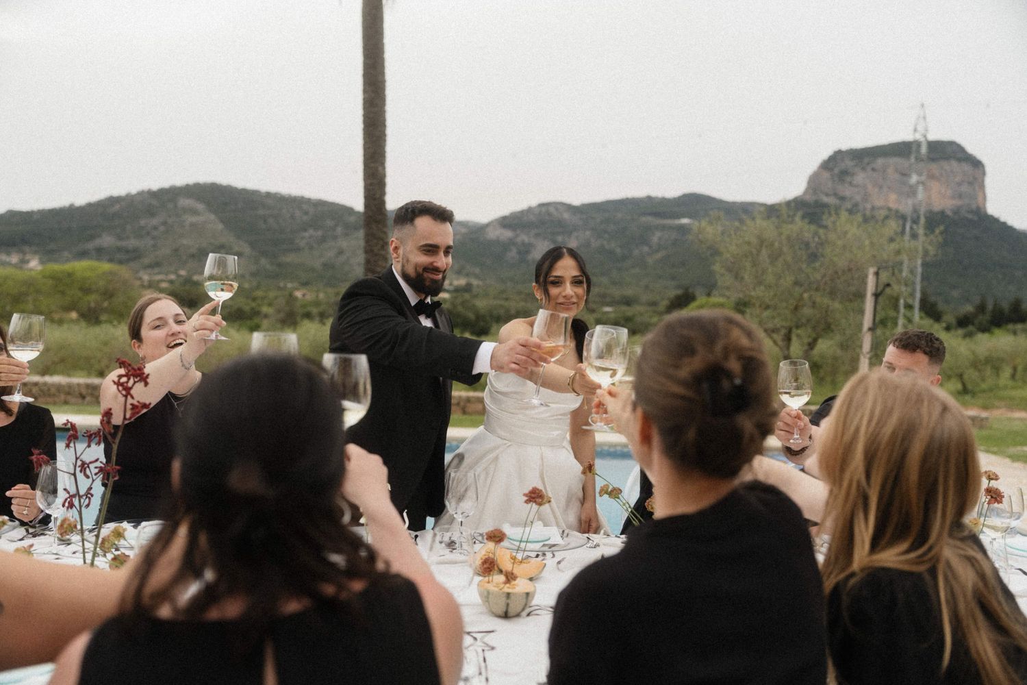 Bride and groom toast with guests at elegant outdoor wedding reception with mountain scenery.