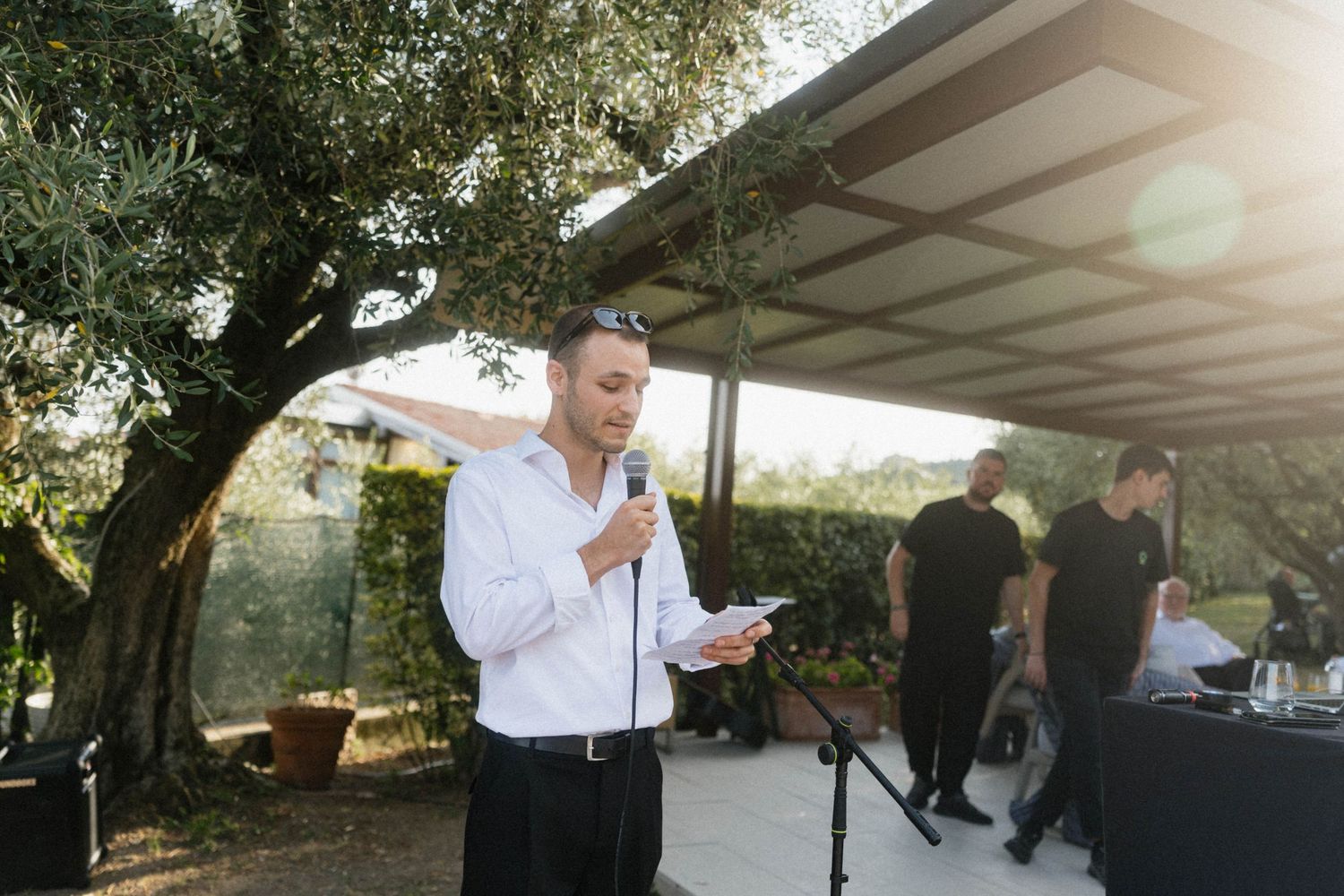 Man in white shirt giving a speech at an outdoor event, holding microphone and paper in sunlit garden.