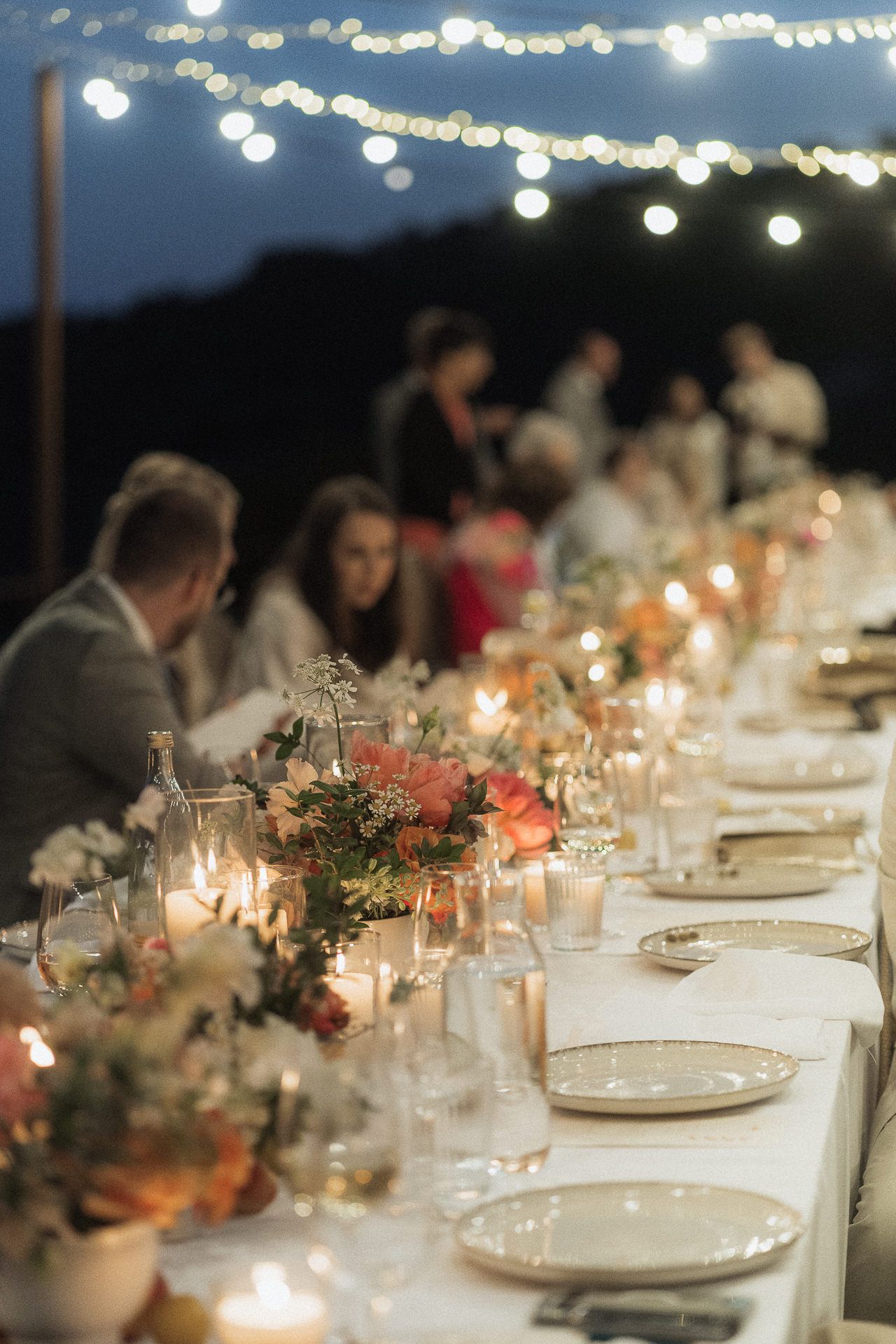 Elegant outdoor wedding reception table with floral centerpieces, candles, and string lights at dusk.