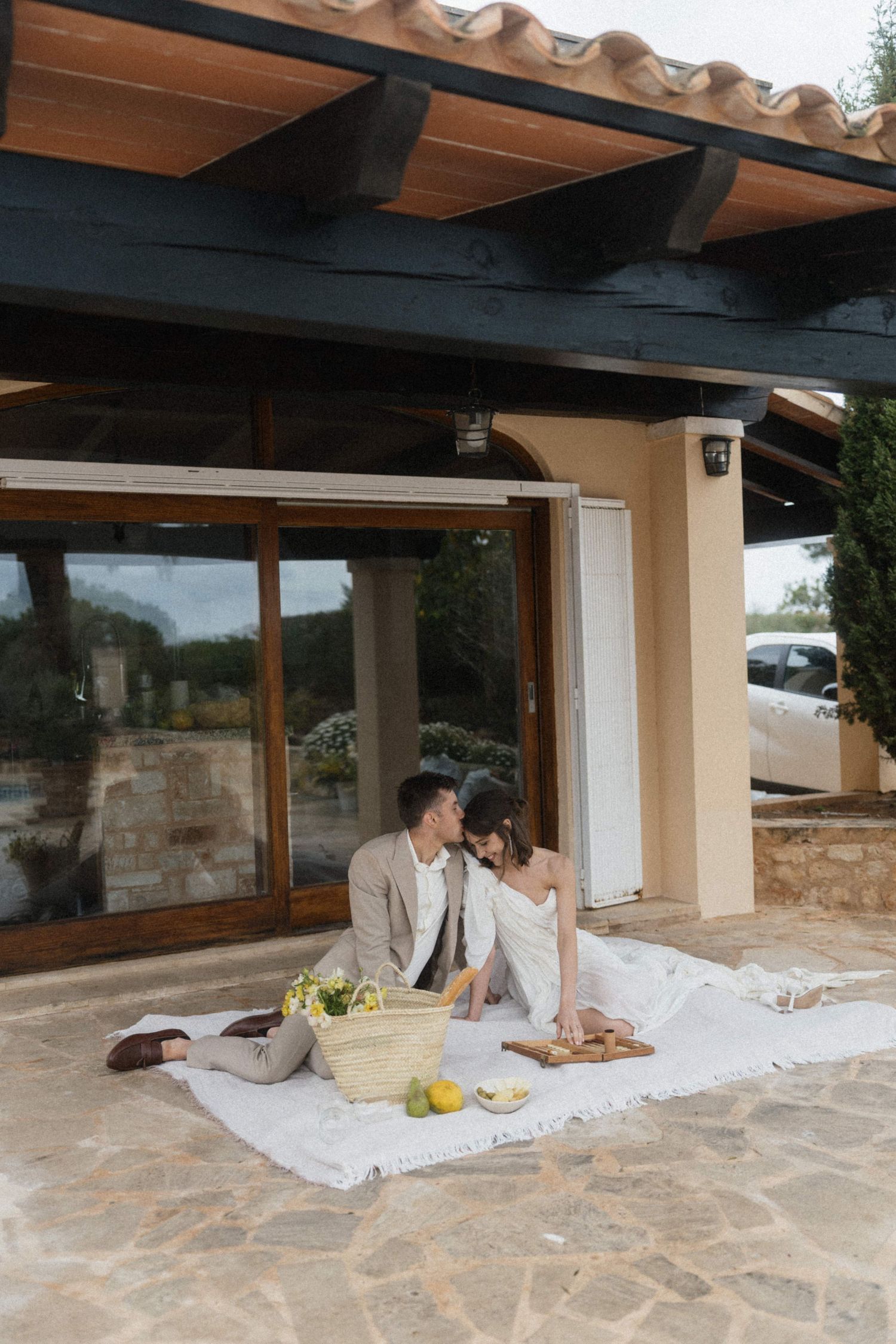 Newlyweds enjoying outdoor picnic on white blanket with fruit basket outside rustic villa.