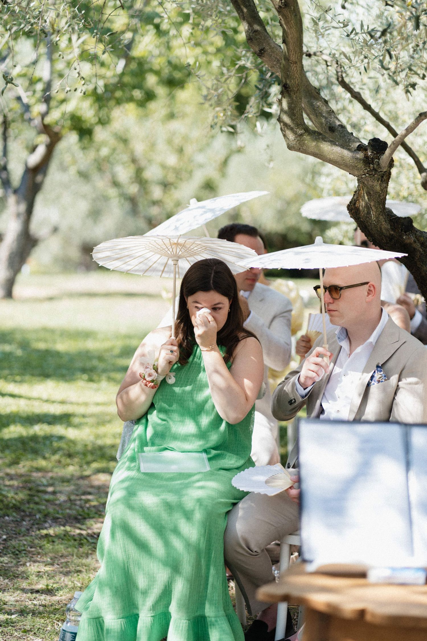 Emotional wedding guest in green dress crying under white parasol during outdoor ceremony in sunny garden.