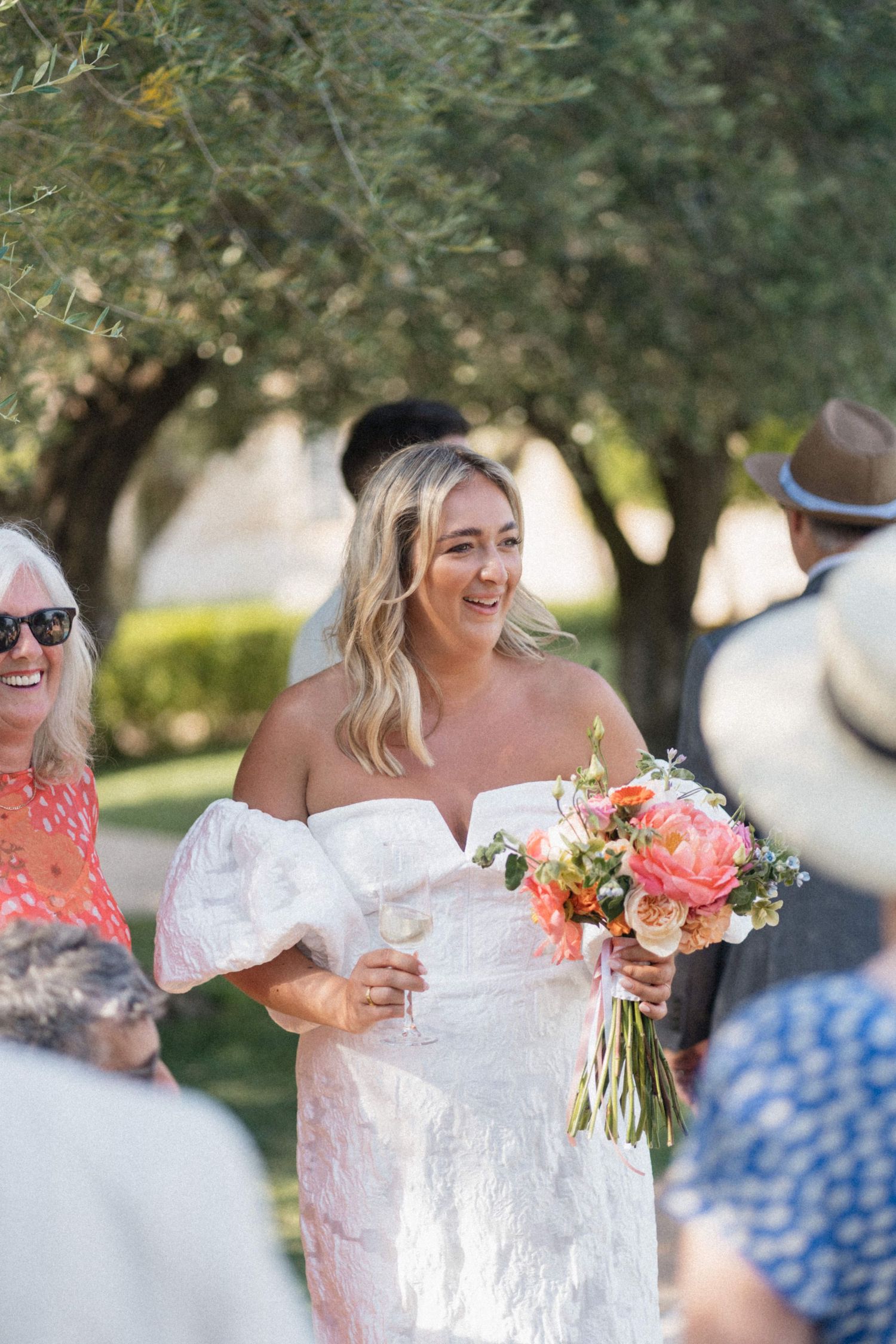 Smiling bride in off-shoulder white gown holding a coral peony bouquet at outdoor wedding.