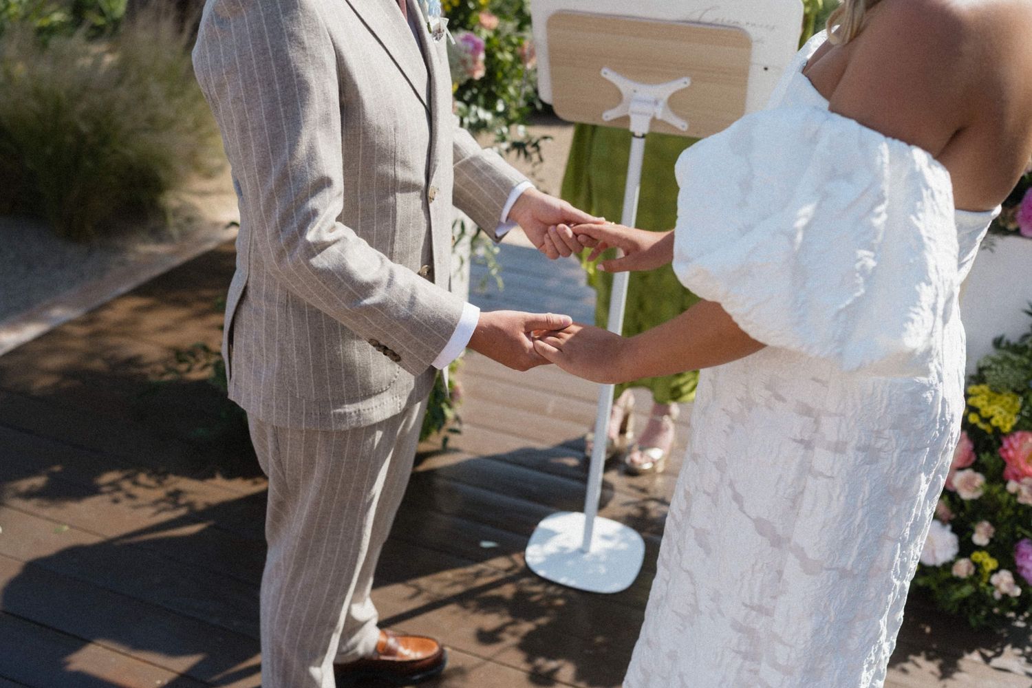 Bride and groom exchanging rings during outdoor wedding ceremony, bride in white lace dress, groom in gray pinstripe suit.