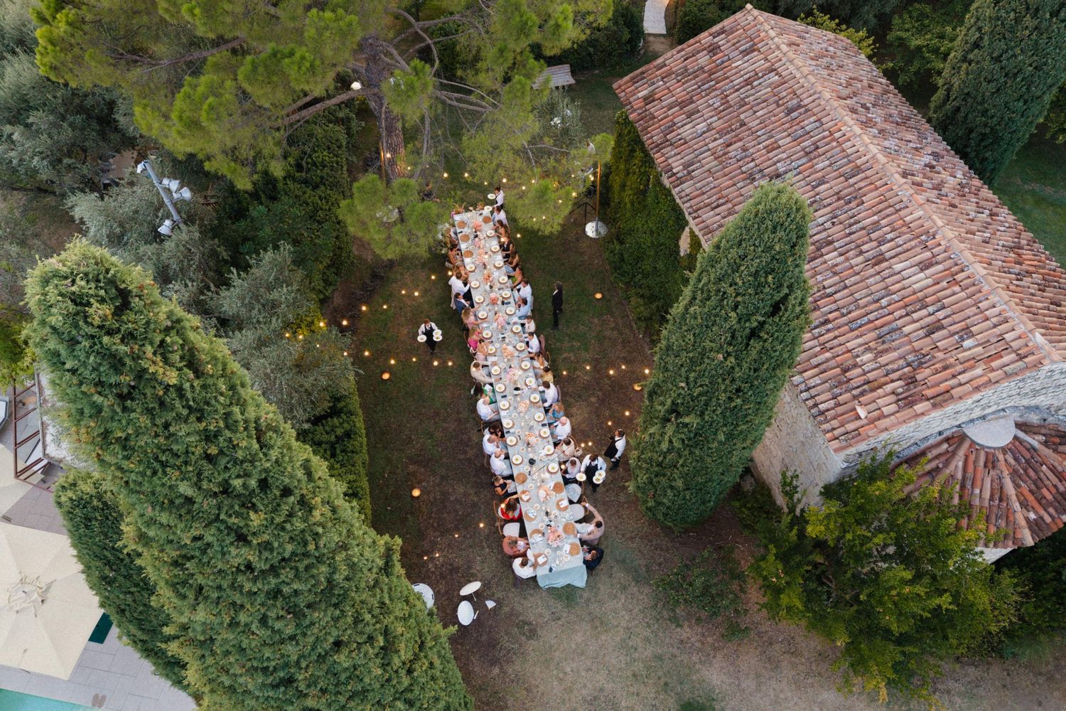 Aerial view of an elegant outdoor wedding reception with long tables set among cypress trees beside a rustic villa.