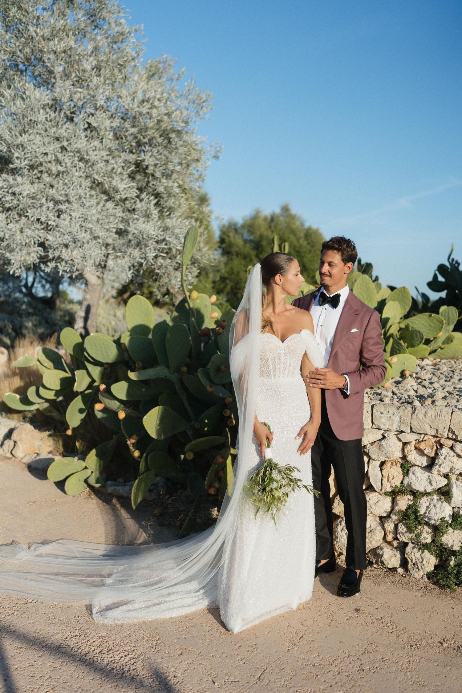 A bride in a lace gown with a long veil holds a bouquet beside her groom in a burgundy blazer and bow tie near prickly pear cacti.