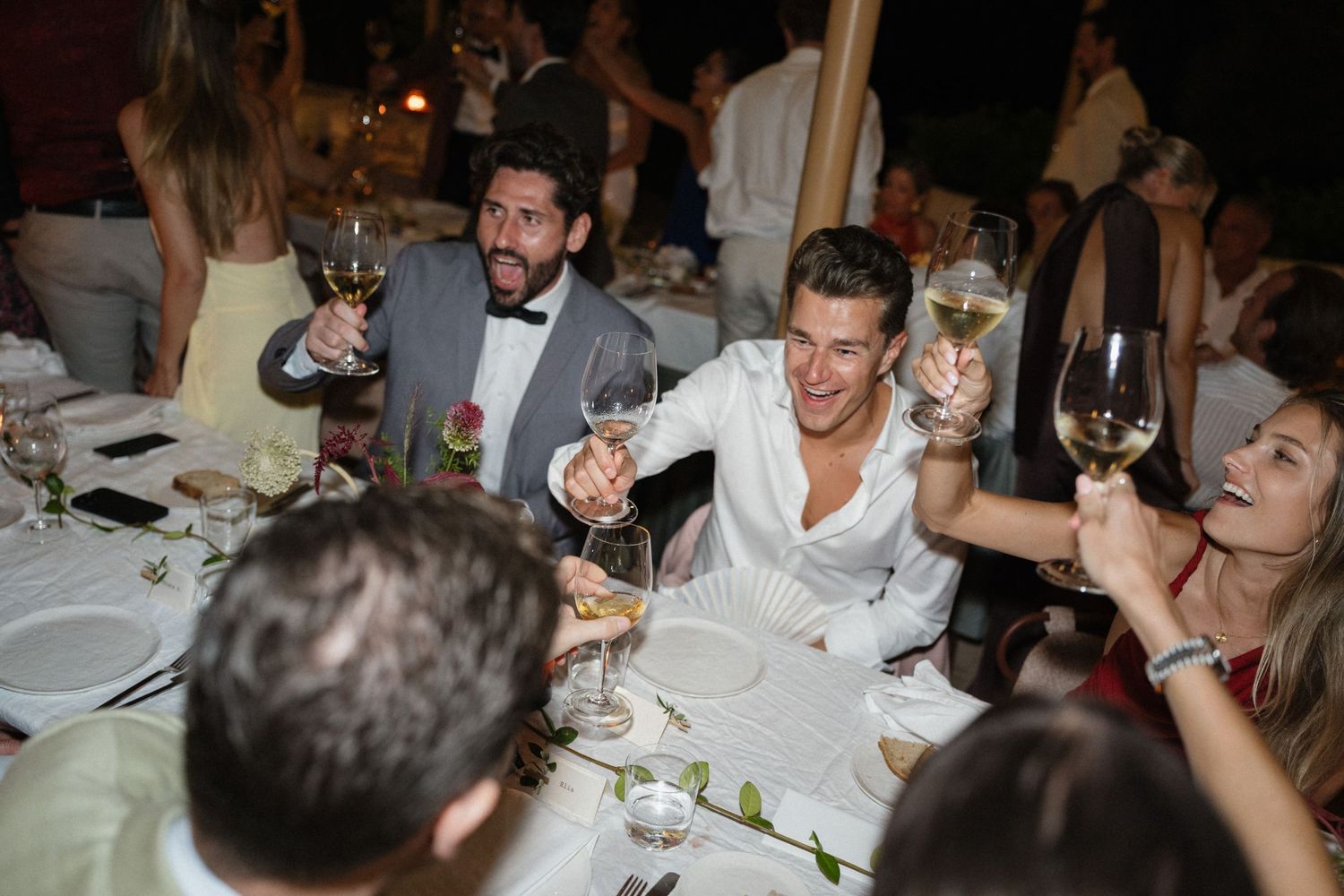 Guests raise wine glasses in a toast at a candlelit dinner table, smiling and celebrating together at an evening event outdoors.