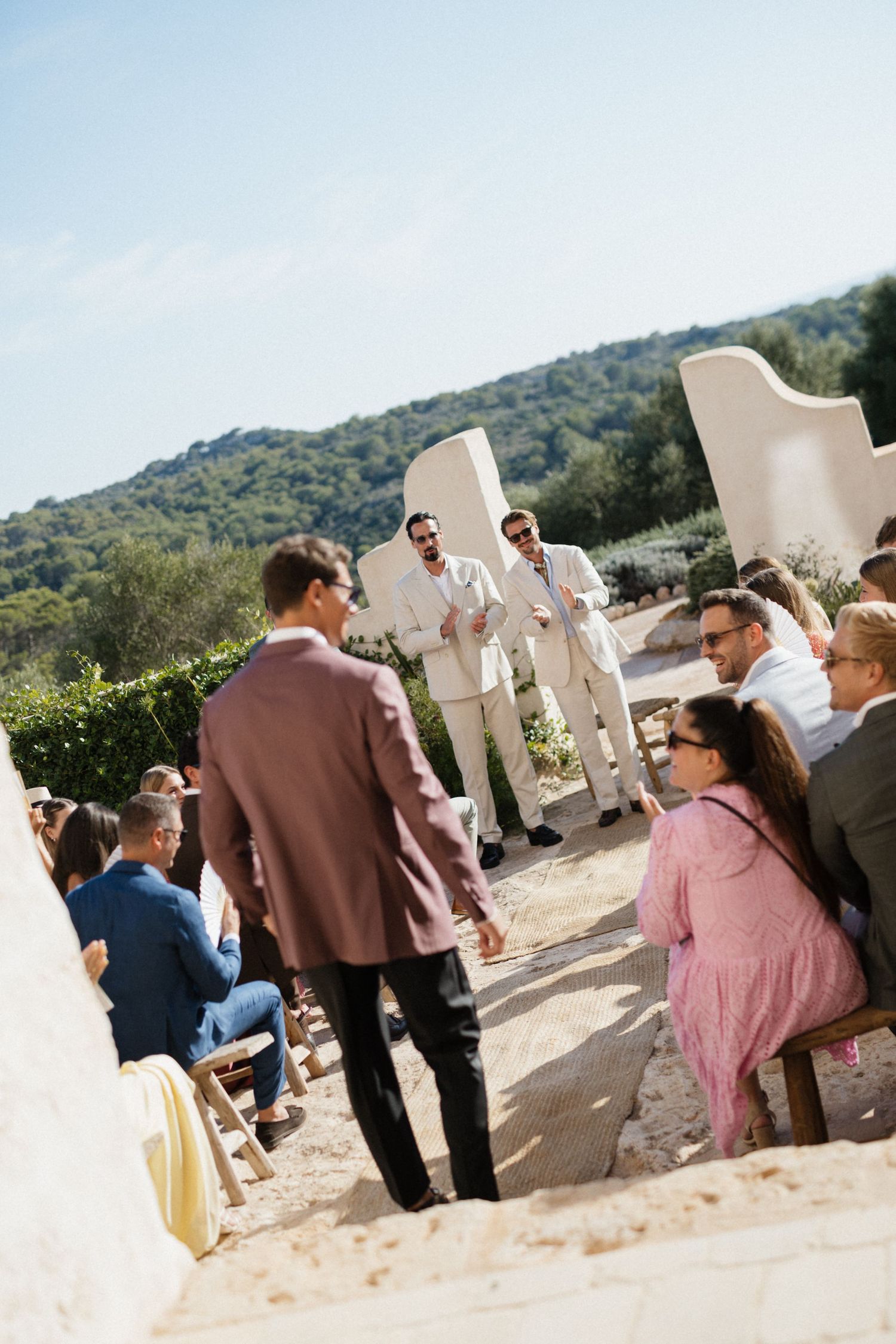 Groomsmen in light suits wait at the end of a sunlit aisle as seated guests look on at a hilltop outdoor wedding ceremony.