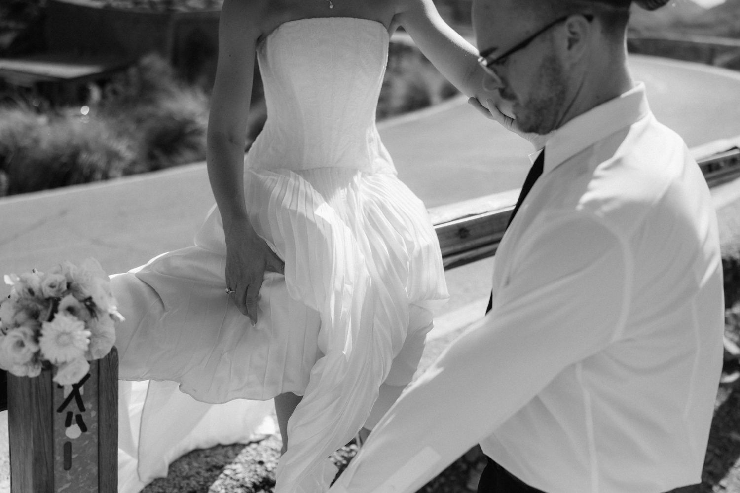Black and white wedding photo of a bride in a flowing gown seated on a boat edge beside a groom with glasses and a bouquet.