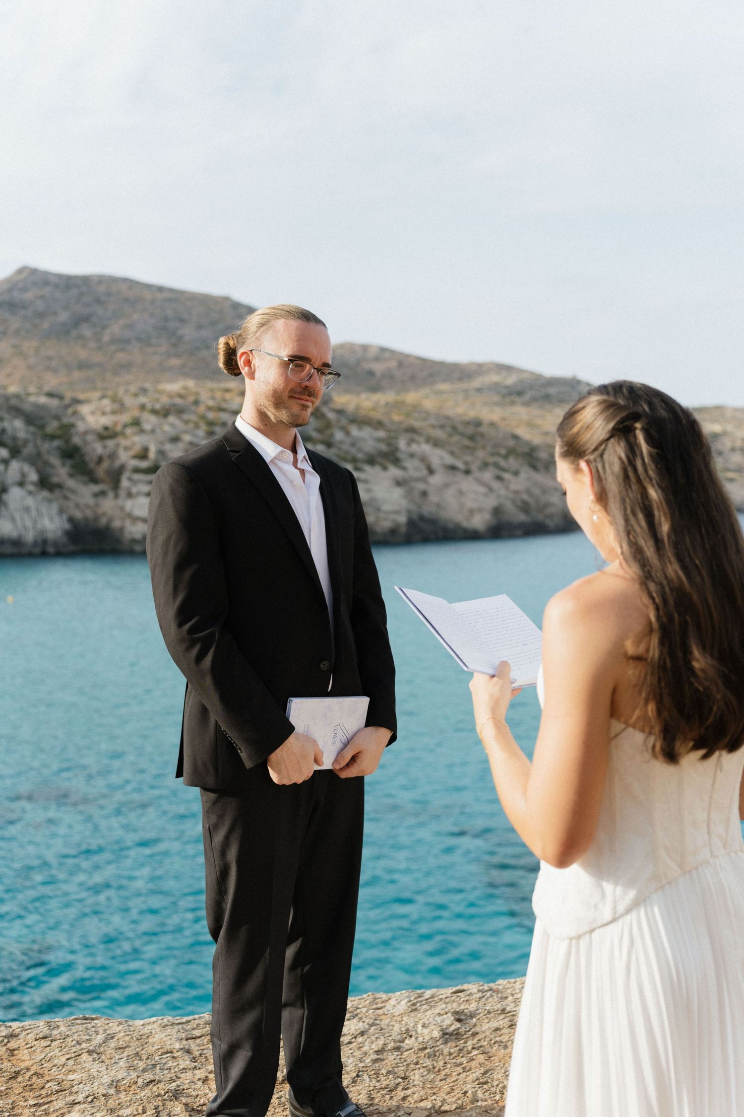 Groom in a black suit smiles as his bride reads her vows during a seaside elopement ceremony overlooking turquoise water and rocky cliffs.