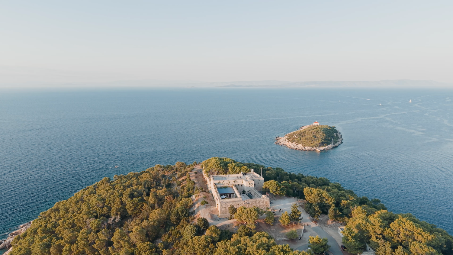 Aerial view of a historic fortress on a lush green peninsula surrounded by calm blue Mediterranean sea waters.