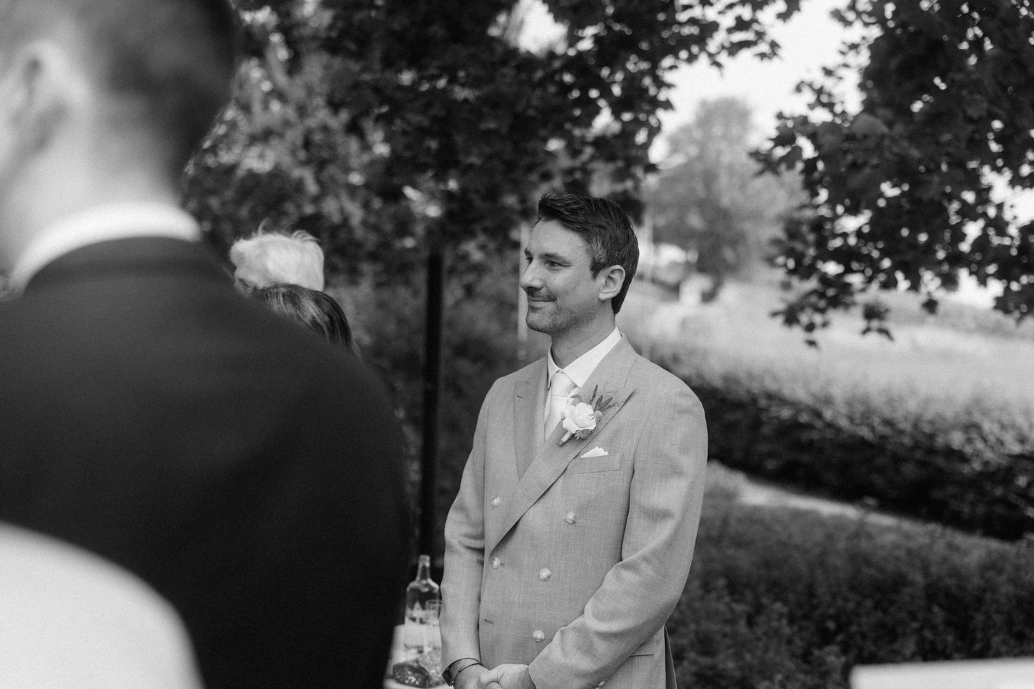 Black and white portrait of smiling groom in boutonniere suit waiting at outdoor wedding ceremony.