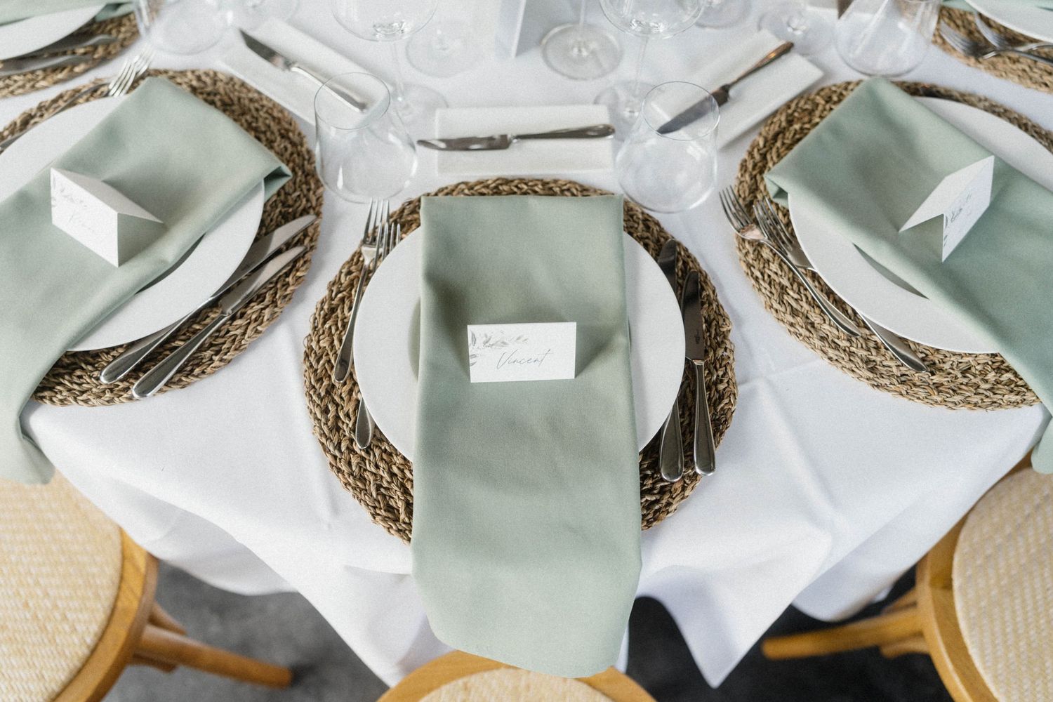 Elegant wedding table setting with sage green napkins, woven chargers, and white place cards.