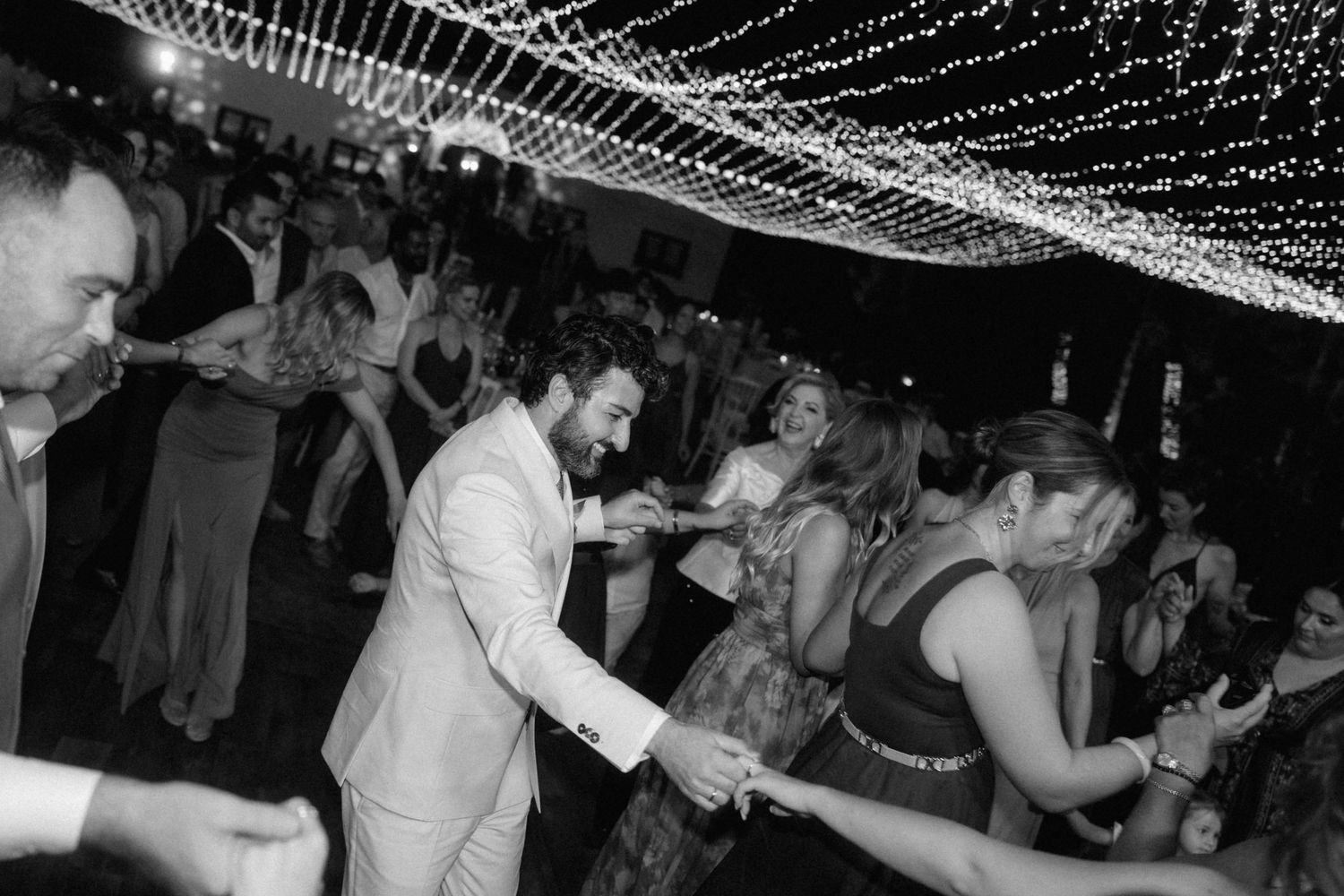 Black and white wedding reception dance floor photo with string lights overhead and guests dancing joyfully.