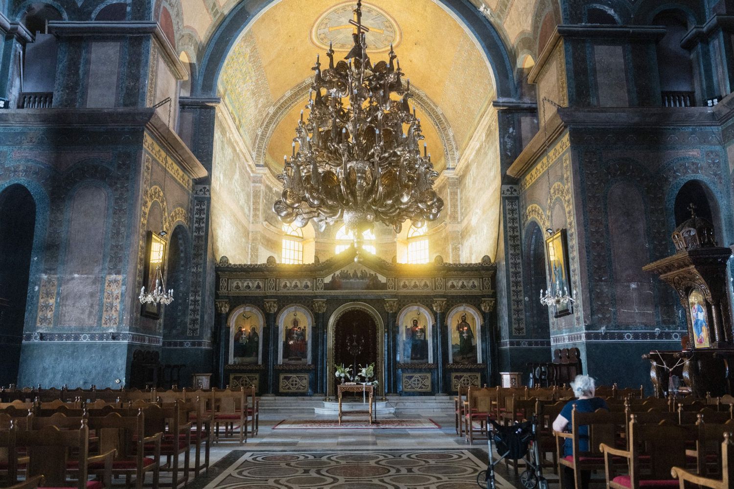 Interior of an ornate Orthodox church featuring a grand chandelier, golden iconostasis, wooden pews, and Byzantine-style architecture.