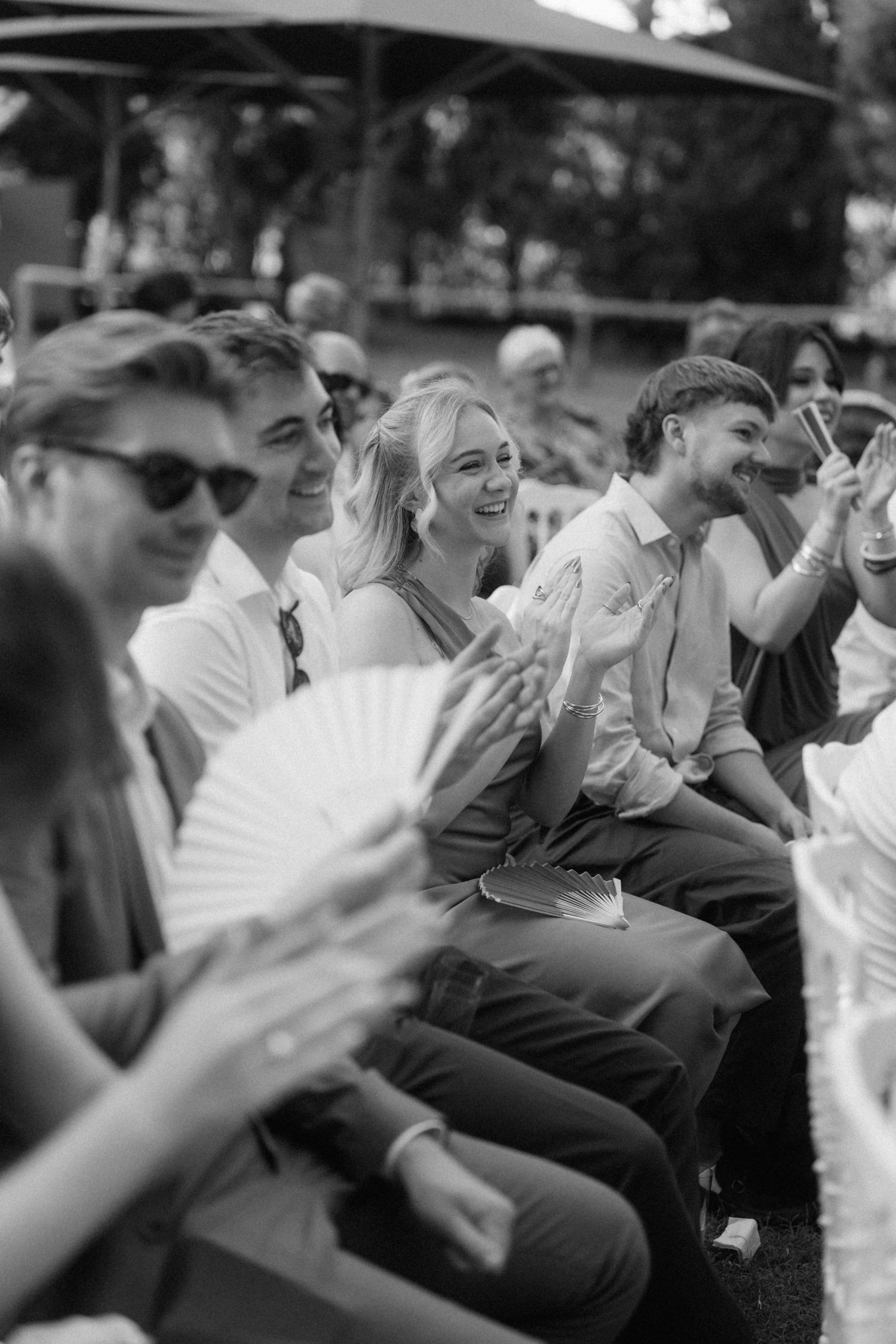 Black and white photo of joyful wedding guests laughing and clapping during outdoor ceremony.