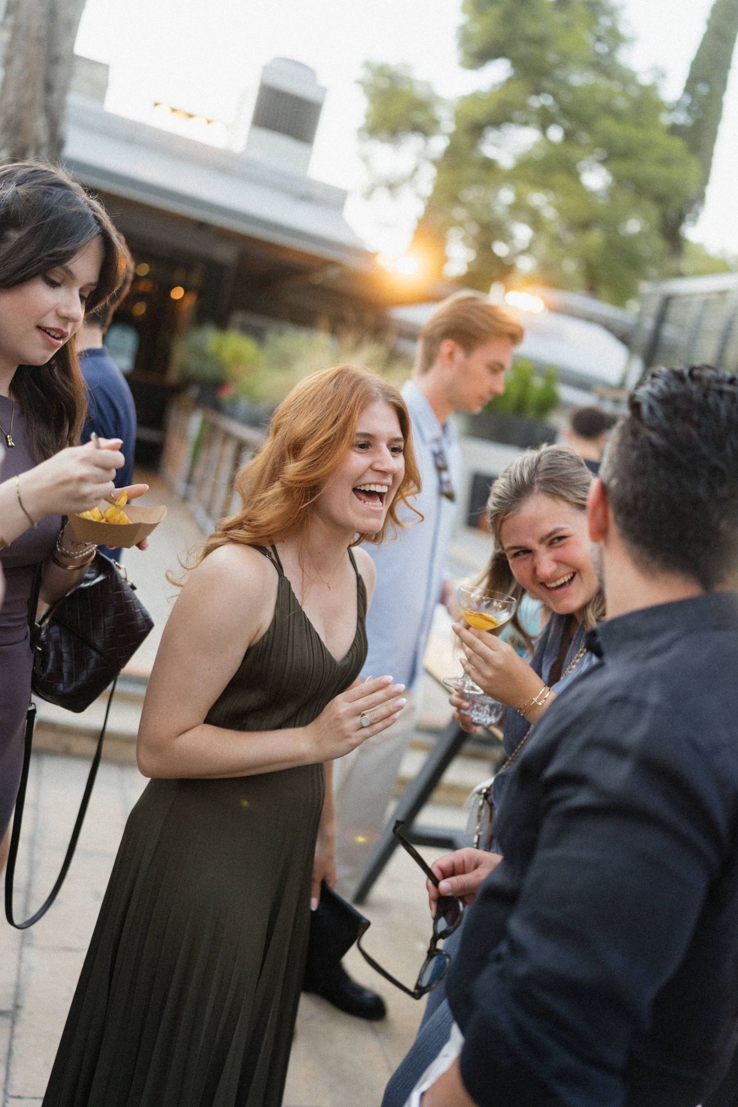 Group of friends laughing and socializing outdoors at sunset event, holding cocktails in elegant evening attire.